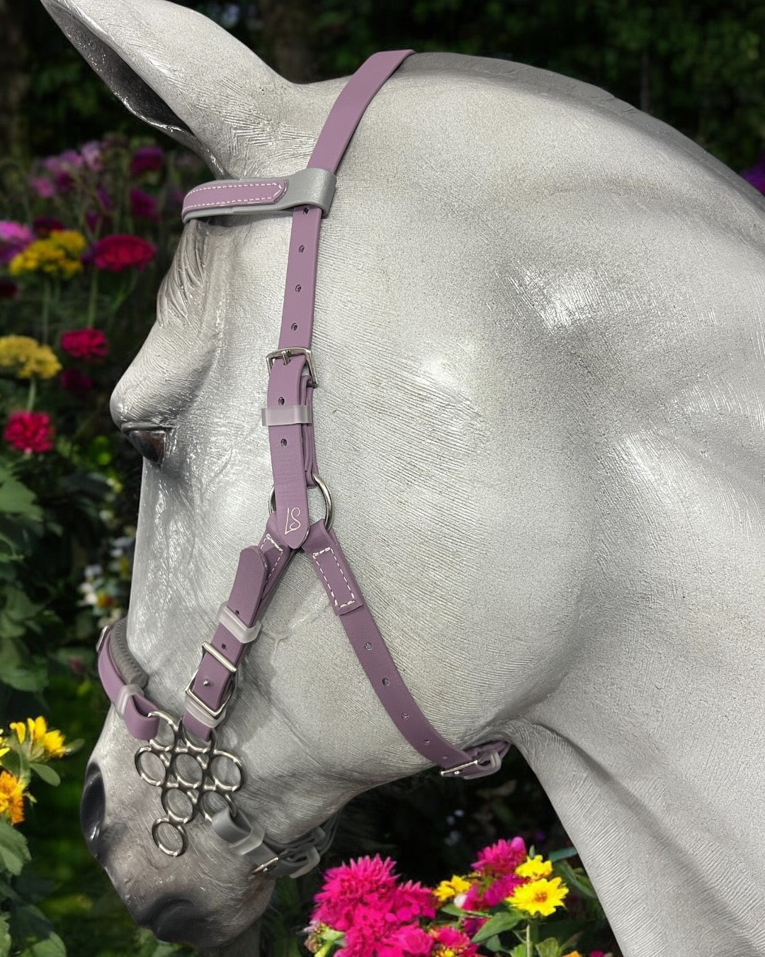 A silver horse with an LS BioThane®️ Hackamore Bridle Package from LS Equestrian, featuring a lavender bridle, stands amid colorful flowers like pink and yellow blooms. The profile close-up highlights its elegant jowl strap against a lush green backdrop.