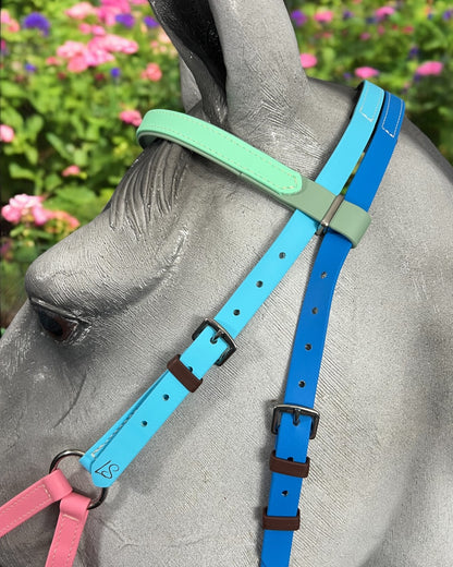 A close-up of a gray horse statue wearing an LS Equestrian "SidePull Bitless Bridle Option 2 - Design Your Own" in blue, green, and pink BioThane®. The background is a blur of vibrant flowers and greenery.