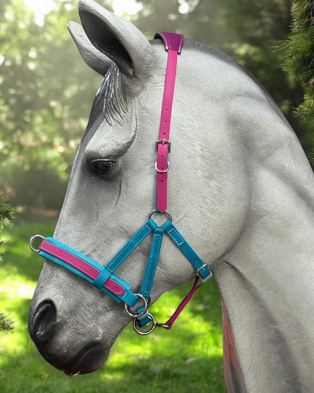 A gray horse stands outdoors in a grassy area with trees in the background, wearing an LS Equestrian Multi Bridle Option 3 - Design your Own BioThane halter, designed with vibrant pink and blue straps.