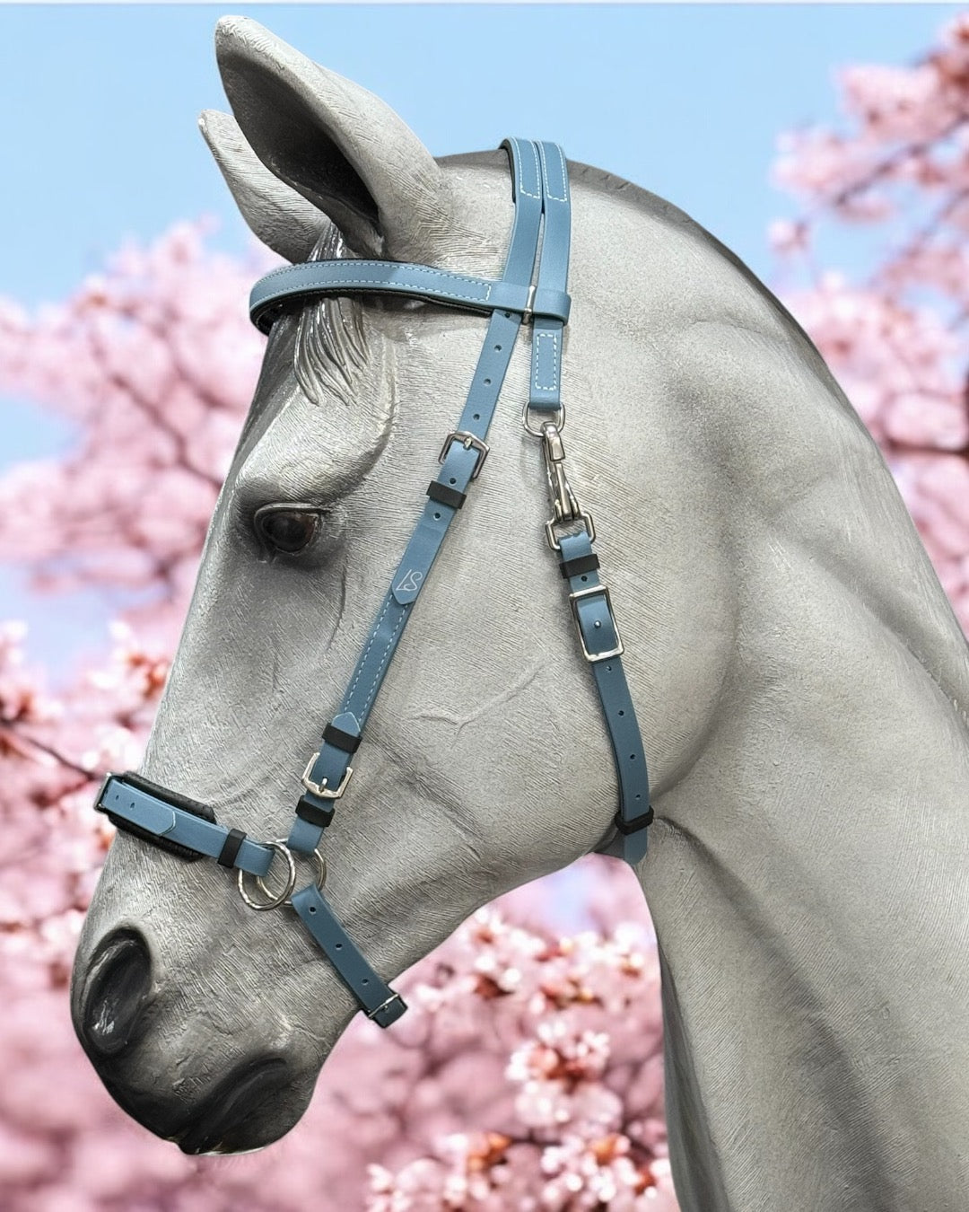 A gray horse statue adorned with a Petrol Blue LS Convertible SidePull / Hackamore Bridle from LS Equestrian NZ stands against a backdrop of pink cherry blossoms. The contrast accentuates the intricate details of the bridle and the delicate flowers, creating a striking visual.