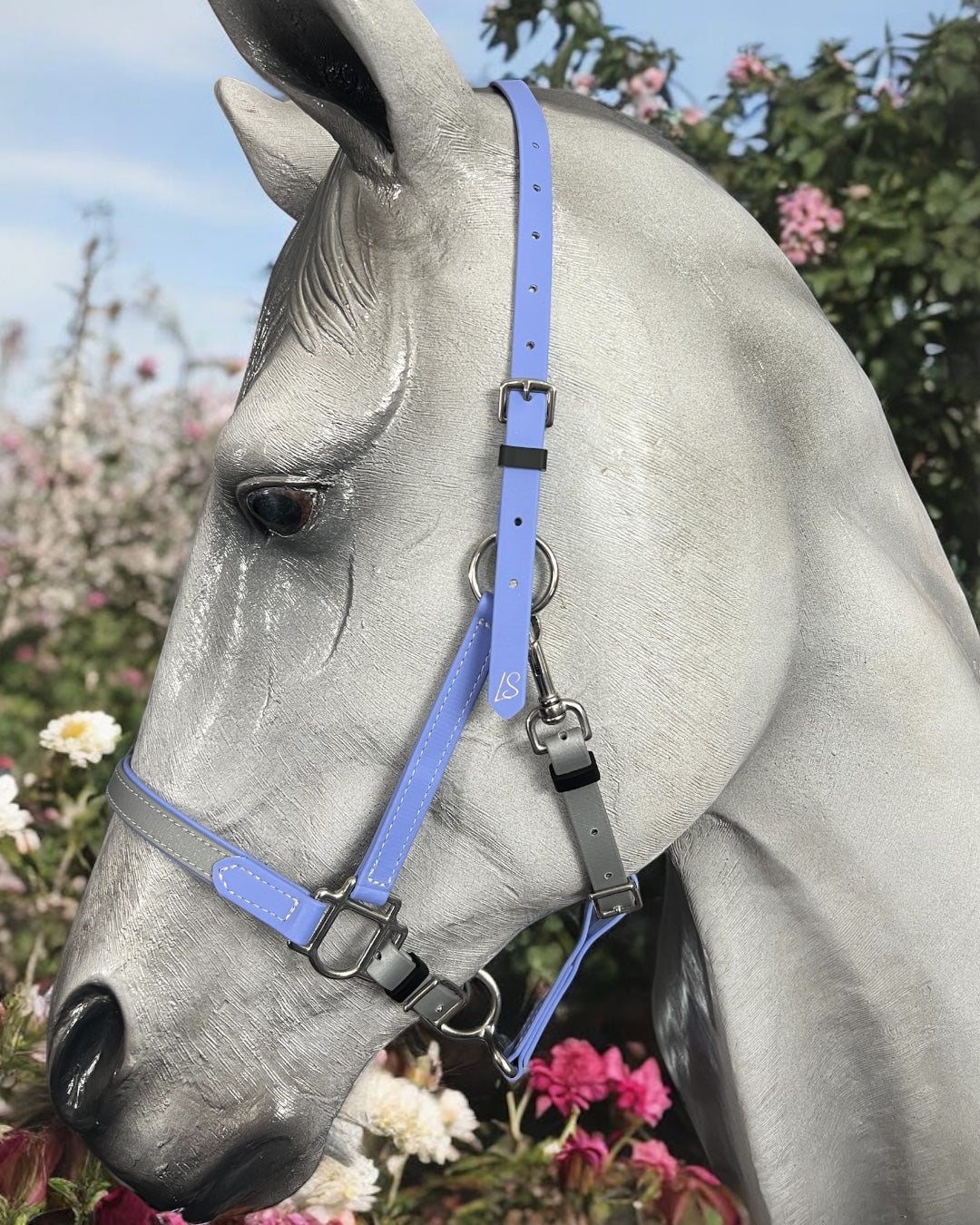 Amidst blooming flowers of pink and white, set against lush green foliage and a clear sky, stands a gray horse donning an LS Equestrian BioThane®️ Deux 2 Part Bridle - Design your own.