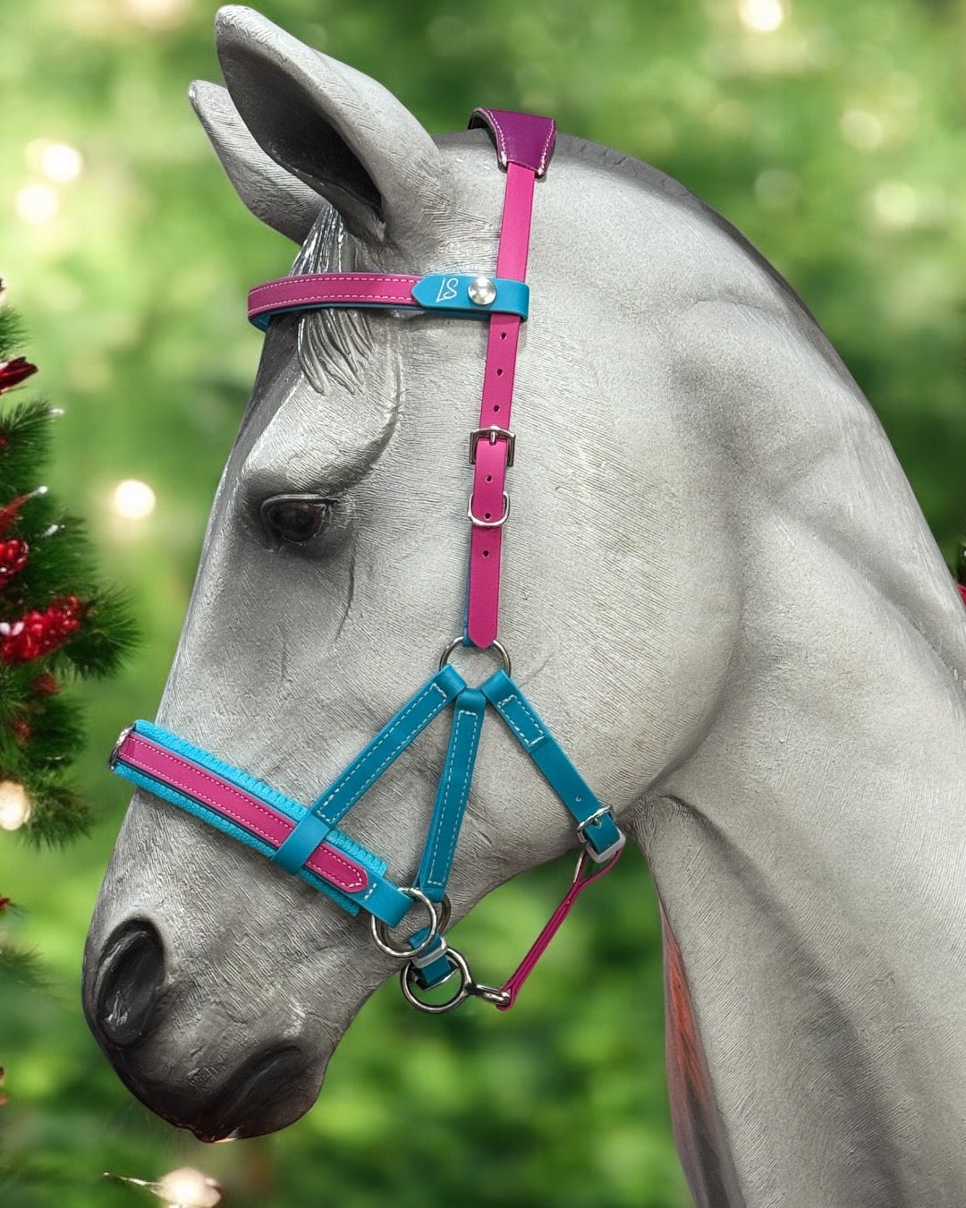 Close-up of a sculpted horse head adorned with the LS Equestrian Multi Bridle Option 3 – Design your Own, featuring colorful BioThane straps in pink and blue leather, complemented by silver buckles. The background is a blur of green foliage accented with hints of red decor, evoking a festive atmosphere.