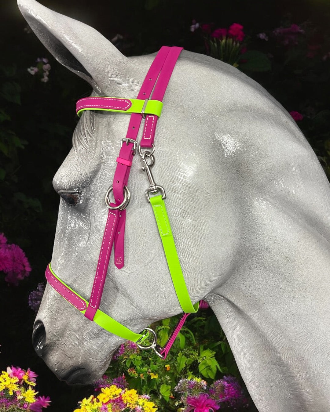 A gray horse wearing an LS Lightweight Enduro Bridle from LS Equestrian NZ in vibrant magenta and apple green stands elegantly against a dark background adorned with colorful flowers.