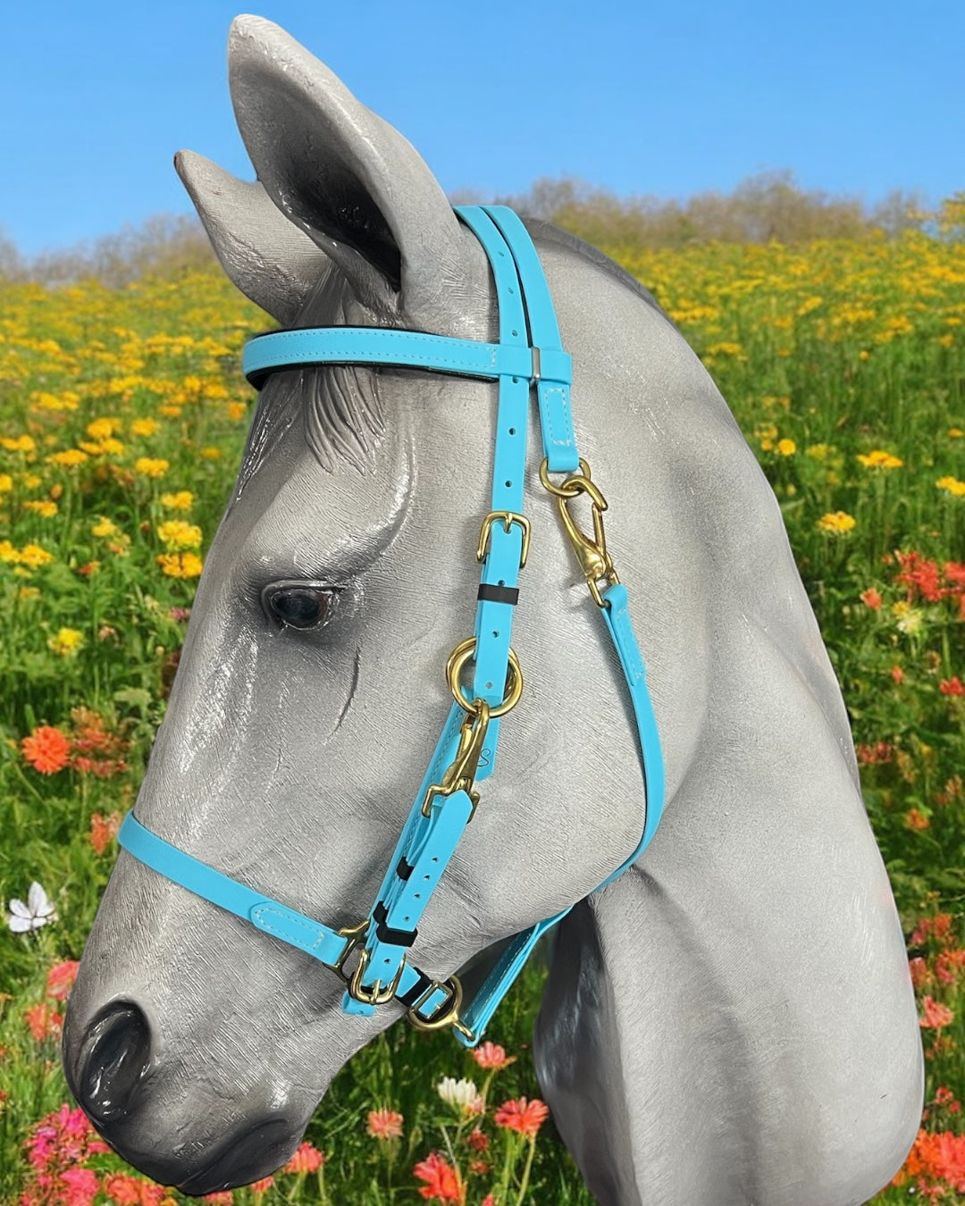 A statue of a horse head showcases a customized LS Enduro bridle from LS Equestrian, featuring gold-colored buckles. The unique design stands out against a backdrop of vibrant yellow and orange flowers beneath a clear blue sky.