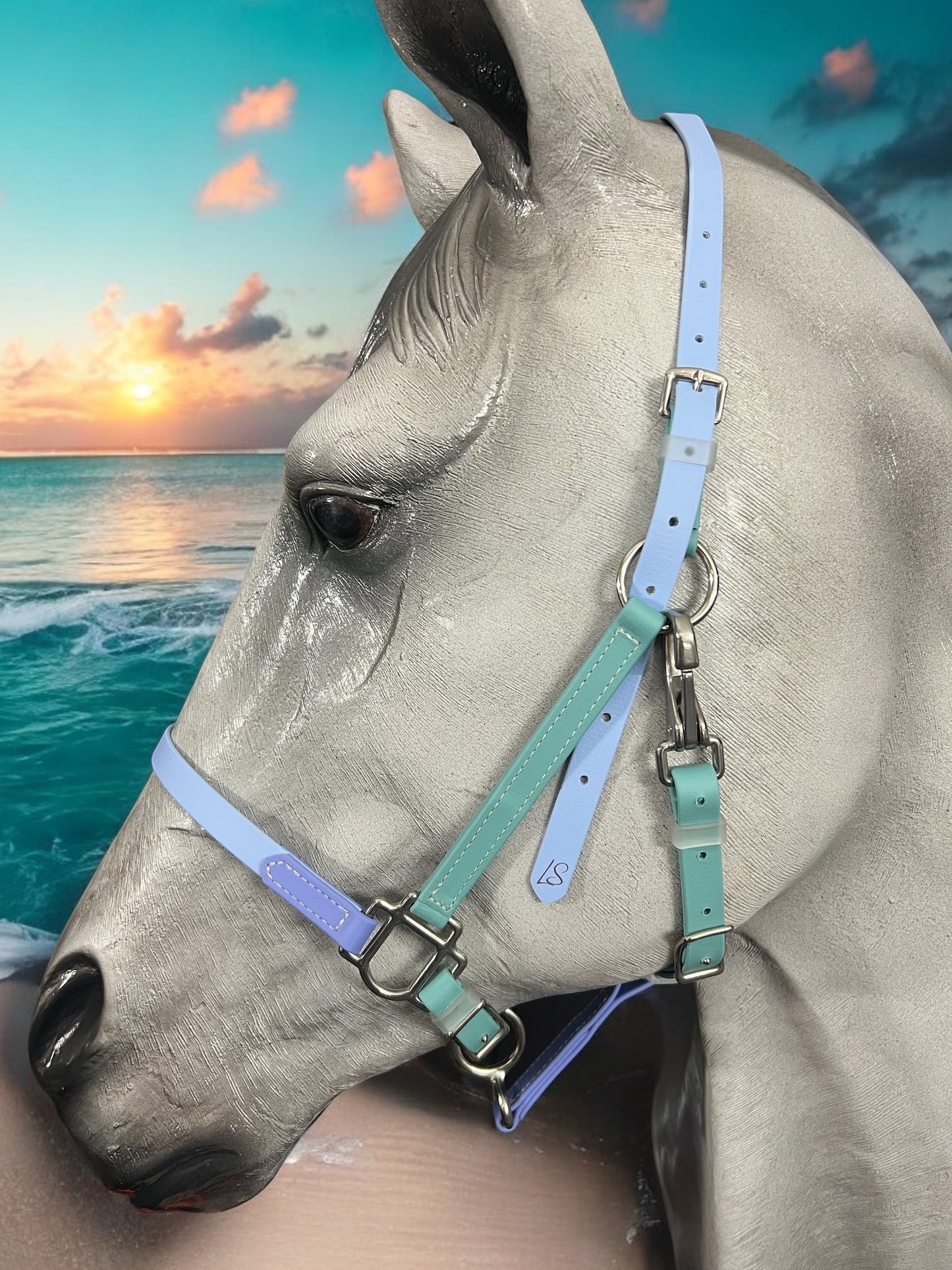 A gray horse, adorned with a pastel blue and green LS BioThane®️ Deux 2 Part Bridle from LS Equestrian, handcrafted in New Zealand, gazes toward the ocean. The background showcases a vibrant sunset with orange and pink hues over the sea.