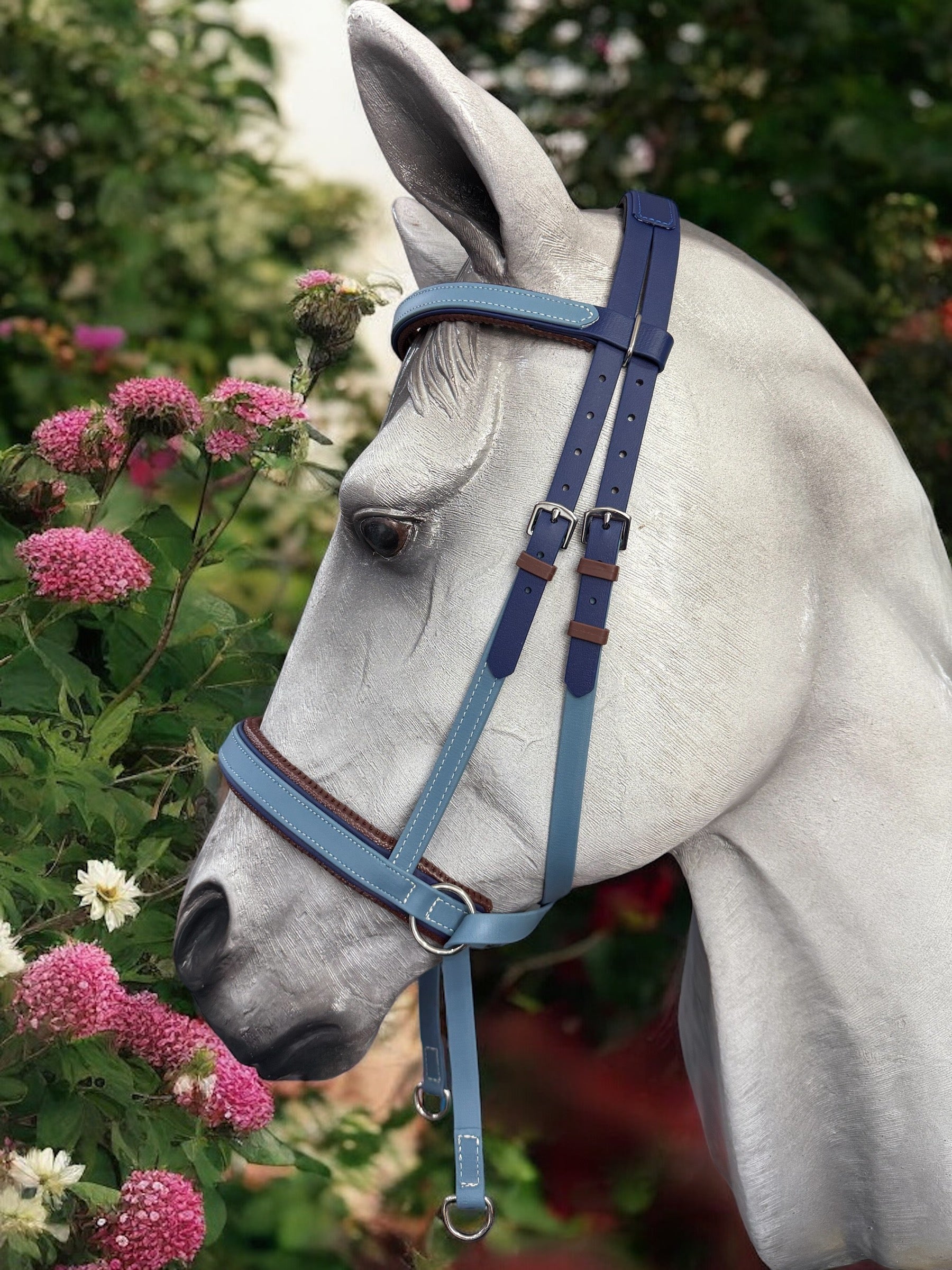 A white horse wearing a Navy & Petrol Blue Cross Under Bitless Bridle from LS Equestrian NZ stands next to a bush adorned with pink and white flowers, set against a backdrop of lush green foliage.