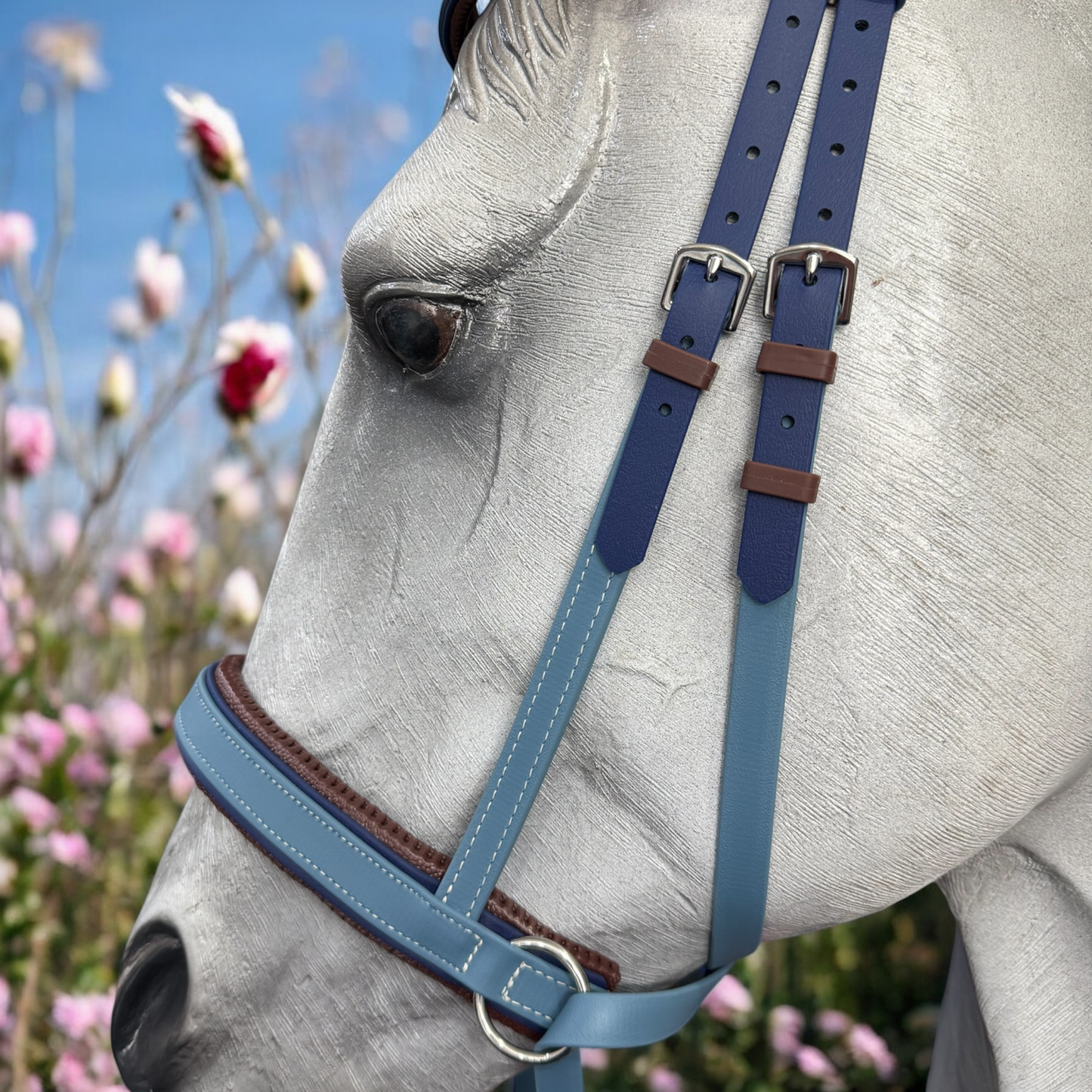 Close-up of a white horse adorned in the LS Equestrian NZ Cross Under Bitless Bridle in Navy & Petrol Blue with white stitching, set against a backdrop of pink and white flowers against a blue sky.