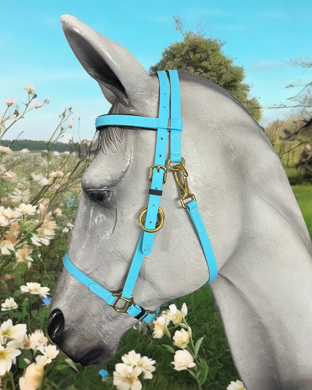 Amid blooming white and pink flowers, a gray horse stands elegantly, adorned in a custom-designed bright blue LS Enduro bridle by LS Equestrian. In the background, lush green trees provide a striking contrast to the clear blue sky.