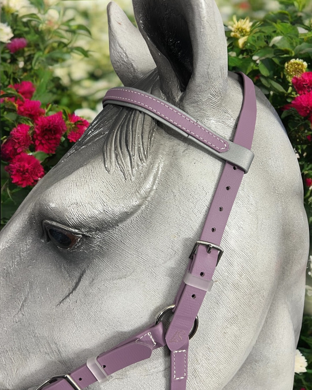 Close-up of a gray horse statue showcases an LS Equestrian design-your-own BioThane®️ Hackamore Bridle Package in purple leather with intricate stitching. Vibrant pink and white flowers surround the statue, forming a colorful and lively background.