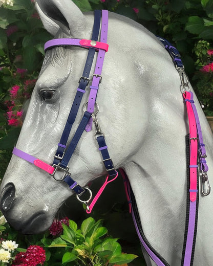 A horse mannequin wears an LS BioThane®️ Deux 2 Part Bridle from LS Equestrian, featuring a personalized design in vibrant shades of pink, purple, and blue. The backdrop displays lush green foliage paired with clusters of pink and white flowers.