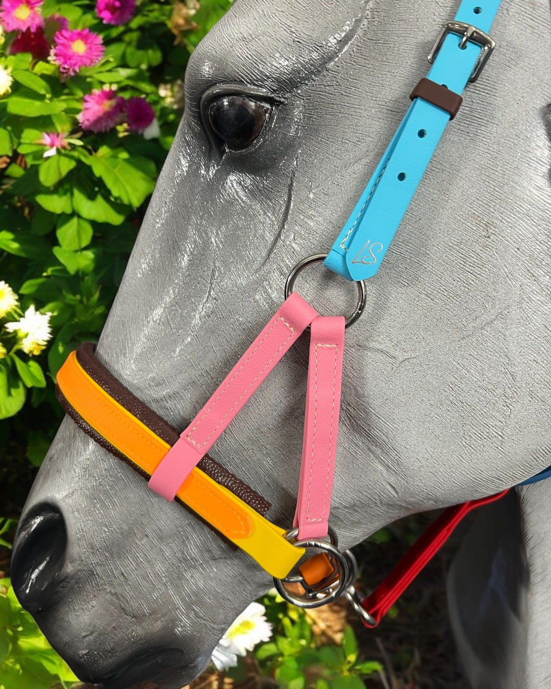 A gray horse statue is adorned with a customizable LS Equestrian SidePull Bitless Bridle Option 2, featuring pink, yellow, and blue straps. It stands amid bright pink and white flowers against a lush green backdrop.