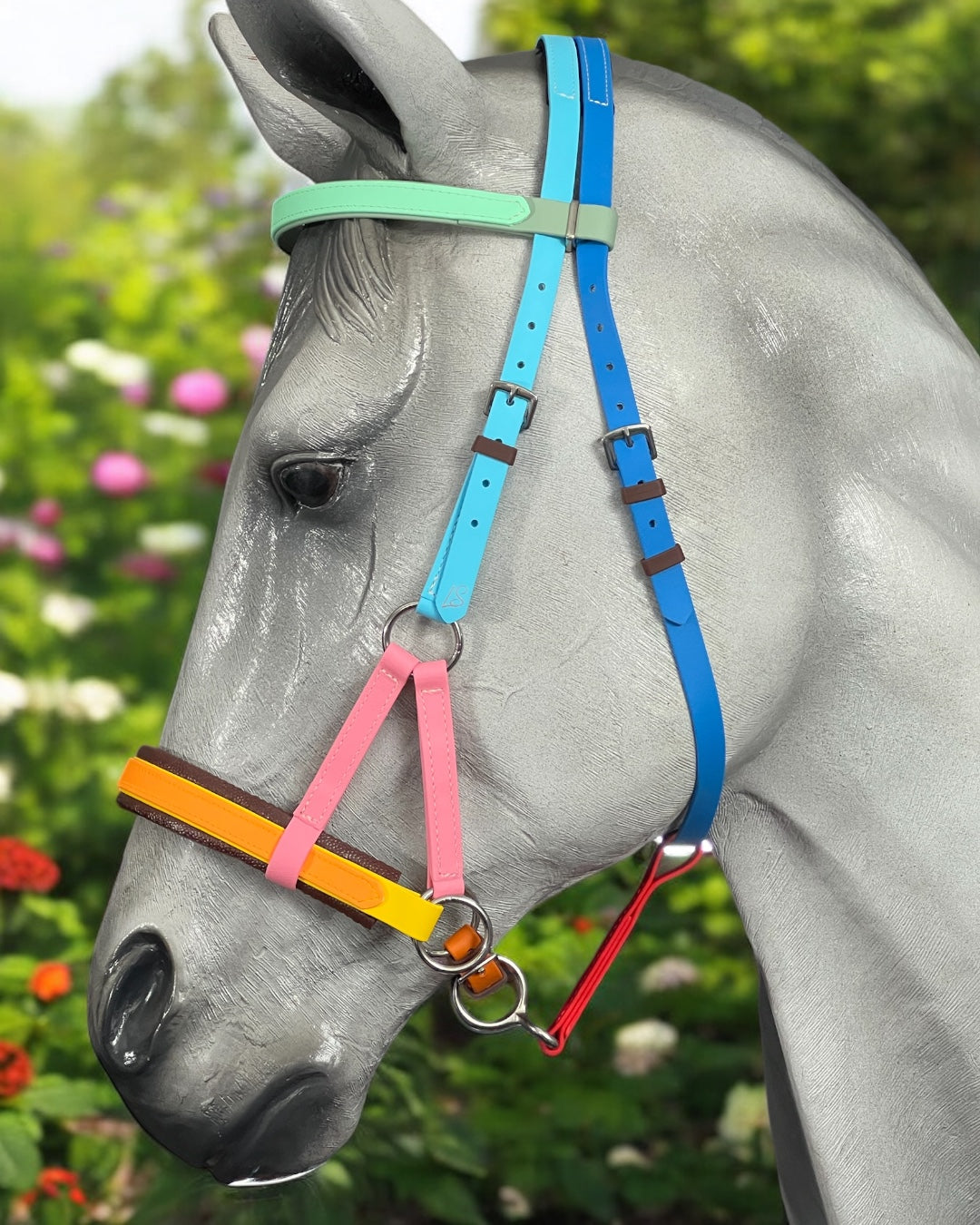 A horse statue wears an LS Equestrian SidePull Bitless Bridle Option 2 with vibrant BioThane®️ straps in blue, green, yellow, pink, and orange. The backdrop features lush green foliage and vibrant red and pink garden flowers.