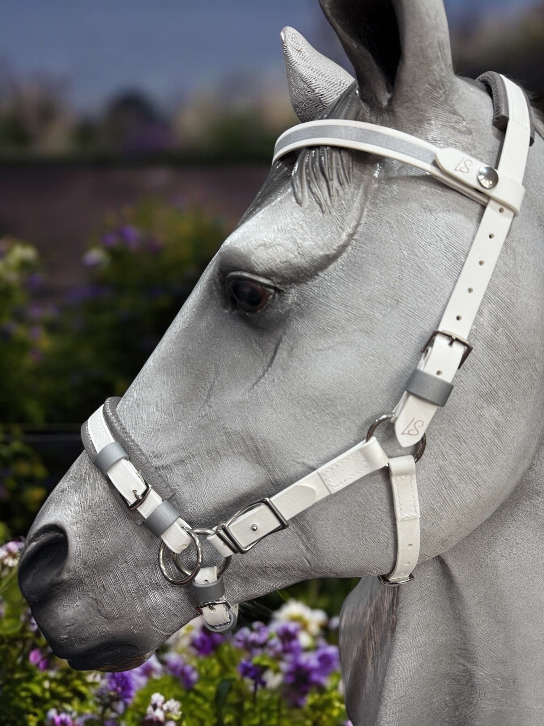 Close-up of a gray horse's head wearing the LS Hackamore Bridle with Jowl Strap by LS Equestrian, set against a background of blurred purple and green flowers, creating a serene and elegant scene.