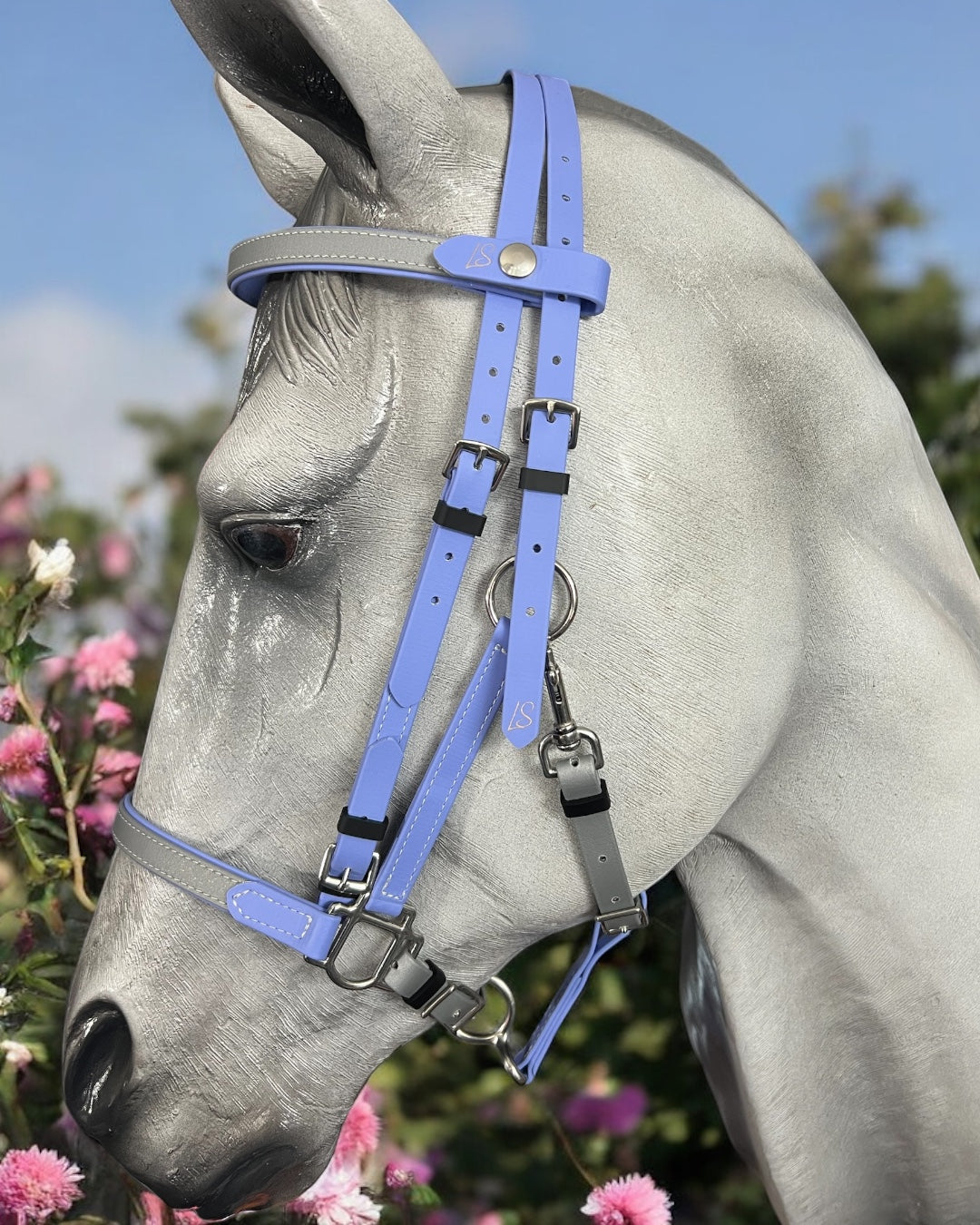 A gray horse is shown in profile against a backdrop of pink flowers and a blue sky, wearing an LS Equestrian bridle. The serene and picturesque scene is completed by the light purple LS BioThane®️ Deux 2 Part Bridle - Design your own. This handmade product from New Zealand features high-quality Beta BioThane craftsmanship.