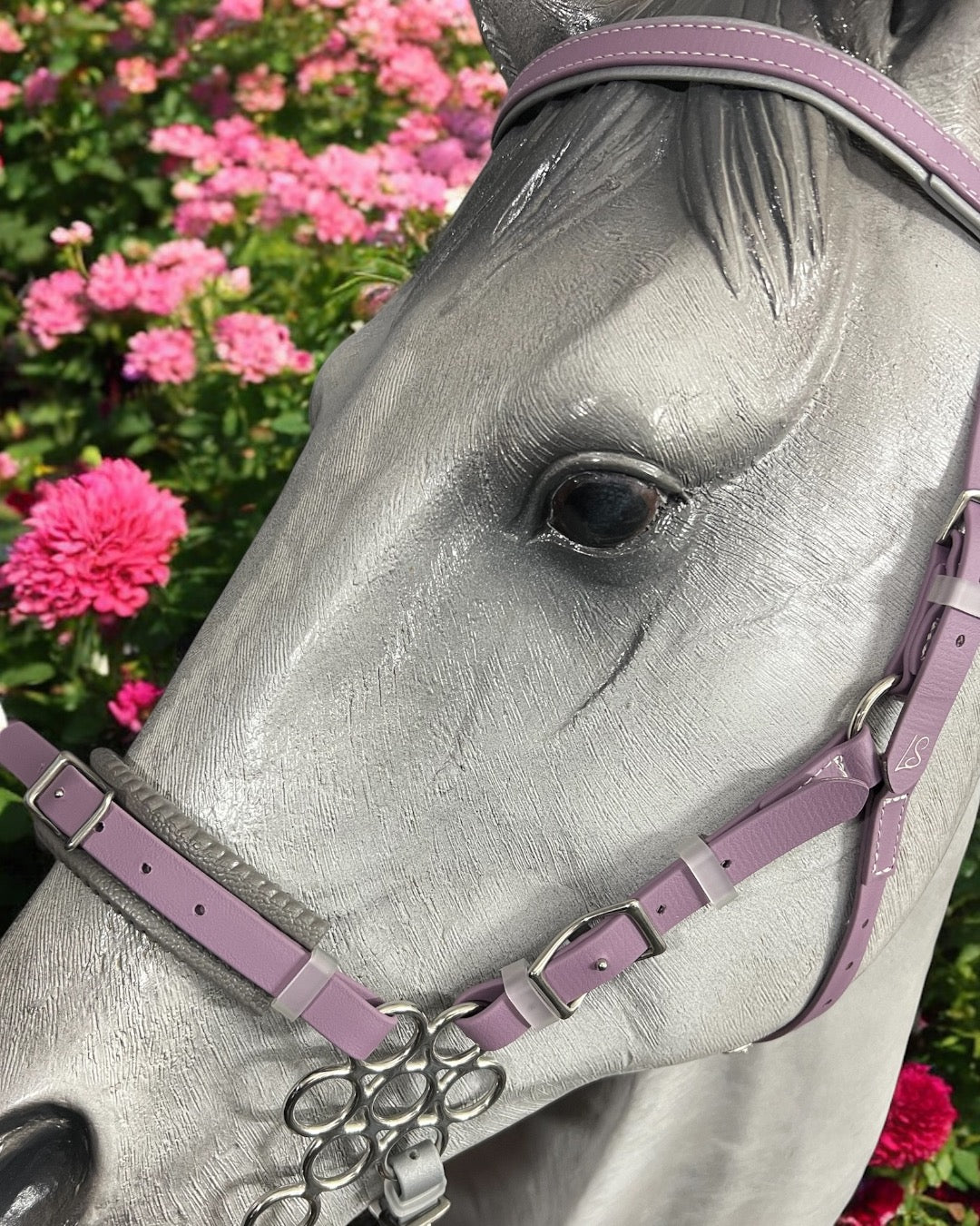 Close-up of a gray horse statue with an LS Hackamore Bridle by LS Equestrian in lavender BioThane®, set against vibrant pink flowers. The realistic texture and gentle expression are highlighted by soft sunlight, while a subtle jowl strap enhances its elegant look.