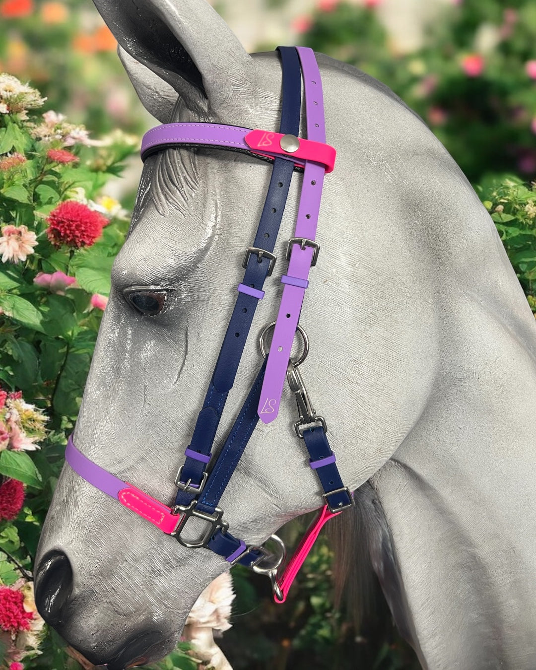 A gray horse wears the vibrant LS BioThane®️ Deux 2 Part Bridle from LS Equestrian, handcrafted in New Zealand with Beta BioThane, showcasing pink, purple, and blue straps amidst blooming red and pink dahlias.