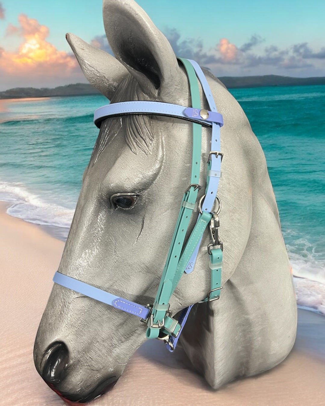 A horse head sculpture, adorned with a pastel blue and mint green LS BioThane®️ Deux 2 Part Bridle by LS Equestrian, handmade in New Zealand, is set against a beach backdrop featuring turquoise water and a blue sky.