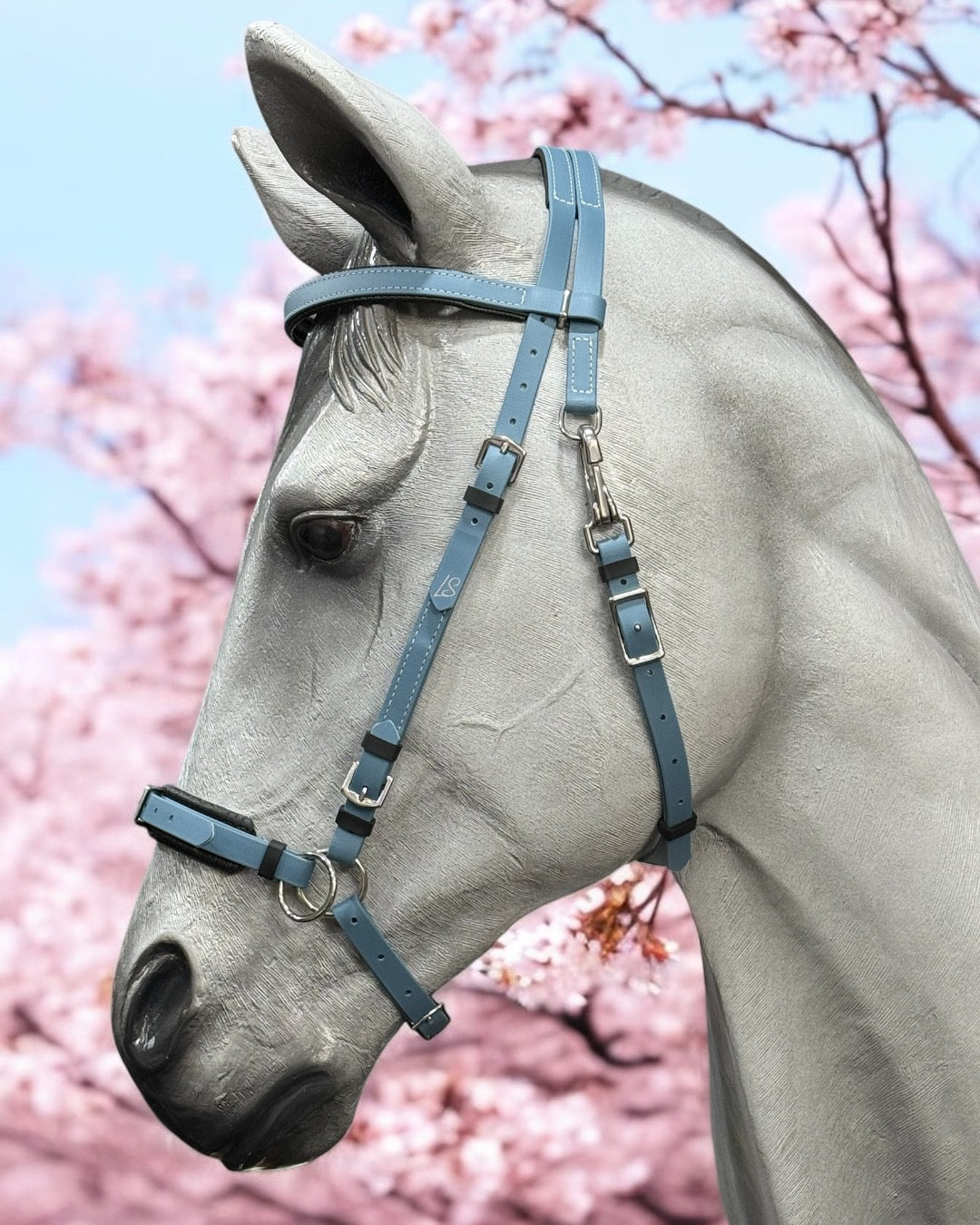 In front of a sea of pink cherry blossoms, a gray horse is adorned with the LS Equestrian NZ's Petrol Blue Convertible SidePull/Hackamore Bridle. The intricately detailed design contrasts beautifully with the soft pastel flowers, showcasing the versatile Beta BioThane material that enhances its multifunctional use.