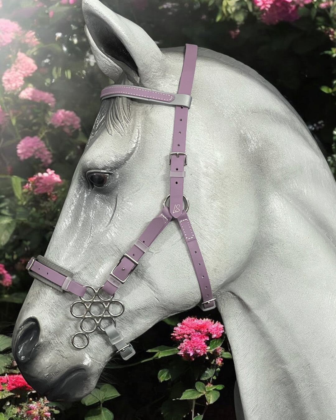 Against a backdrop of pink flowers, a gray horse wears an LS Equestrian BioThane®️ Hackamore Bridle. Its head is in profile, showcasing the bridle's sleek design and purple color.