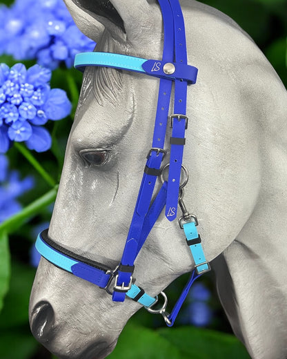 A grey horse with a vibrant blue and teal LS Equestrian Deux 2 Part Bridle stands in front of colorful blue flowers. The bridle, handmade in New Zealand, showcases the brand's logo and includes adjustable straps. The scene is set against a backdrop of lush green leaves.