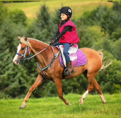 A person wearing a red vest and black helmet rides a brown and white horse with an LS BioThane®️ Deux 2 Part Bridle from LS Equestrian, handmade in New Zealand, and a purple saddle pad across a grassy field. Evergreen trees and a blurred hilly landscape serve as the backdrop.