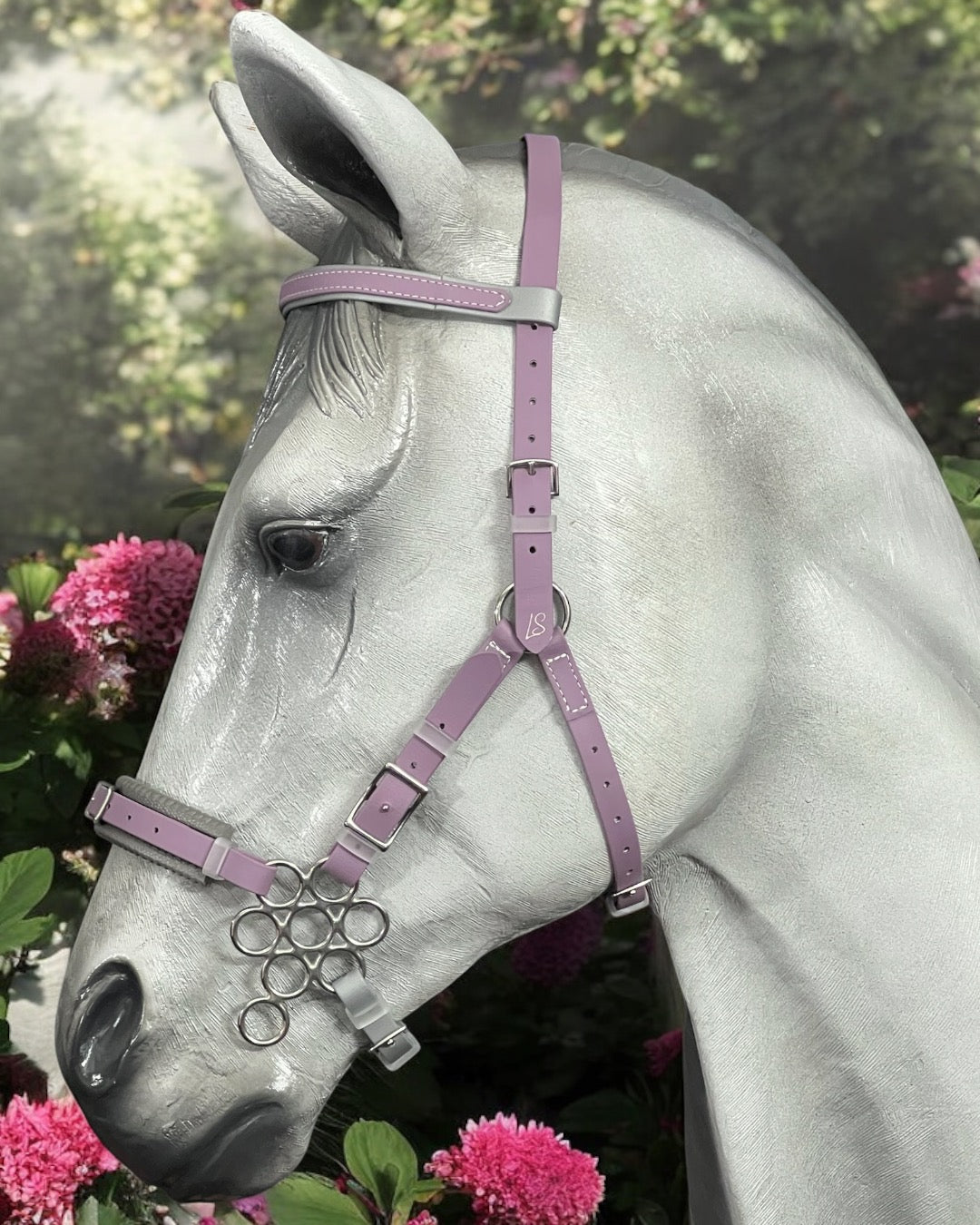 A graceful gray horse, donning an LS Equestrian LS Hackamore Bridle with Jowl Strap in light purple, enhanced with decorative rings and BioThane® artistry, stands amid pink flowers and lush greenery.