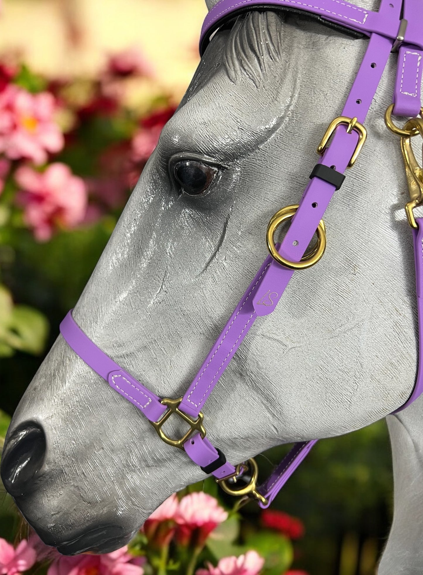 A close-up of a gray horse's head wearing the LS Equestrian LS Enduro - Design your own bridle in vibrant purple leather with brass hardware. Custom-designed details shine against a backdrop of soft-focus pink flowers and greenery, enhancing the scene’s vividness.