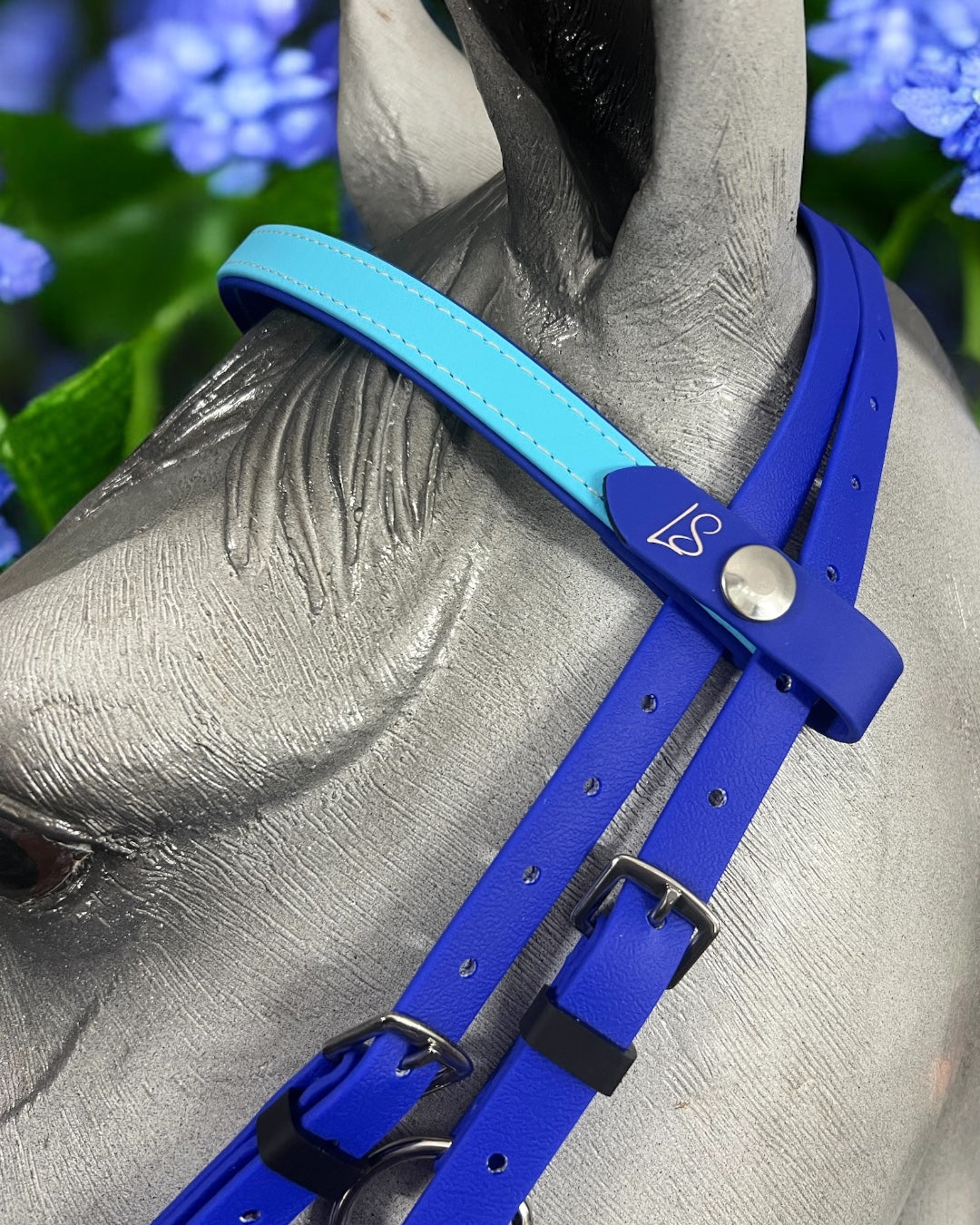 A close-up shot of a horse wearing an LS Equestrian Deux 2 Part Bridle Padded, handcrafted in New Zealand. It features a blue strap made from durable Beta BioThane with silver buckles and proudly showcases the "LS" logo on a silver stud, all set against the soft blur of purple flowers in the background.