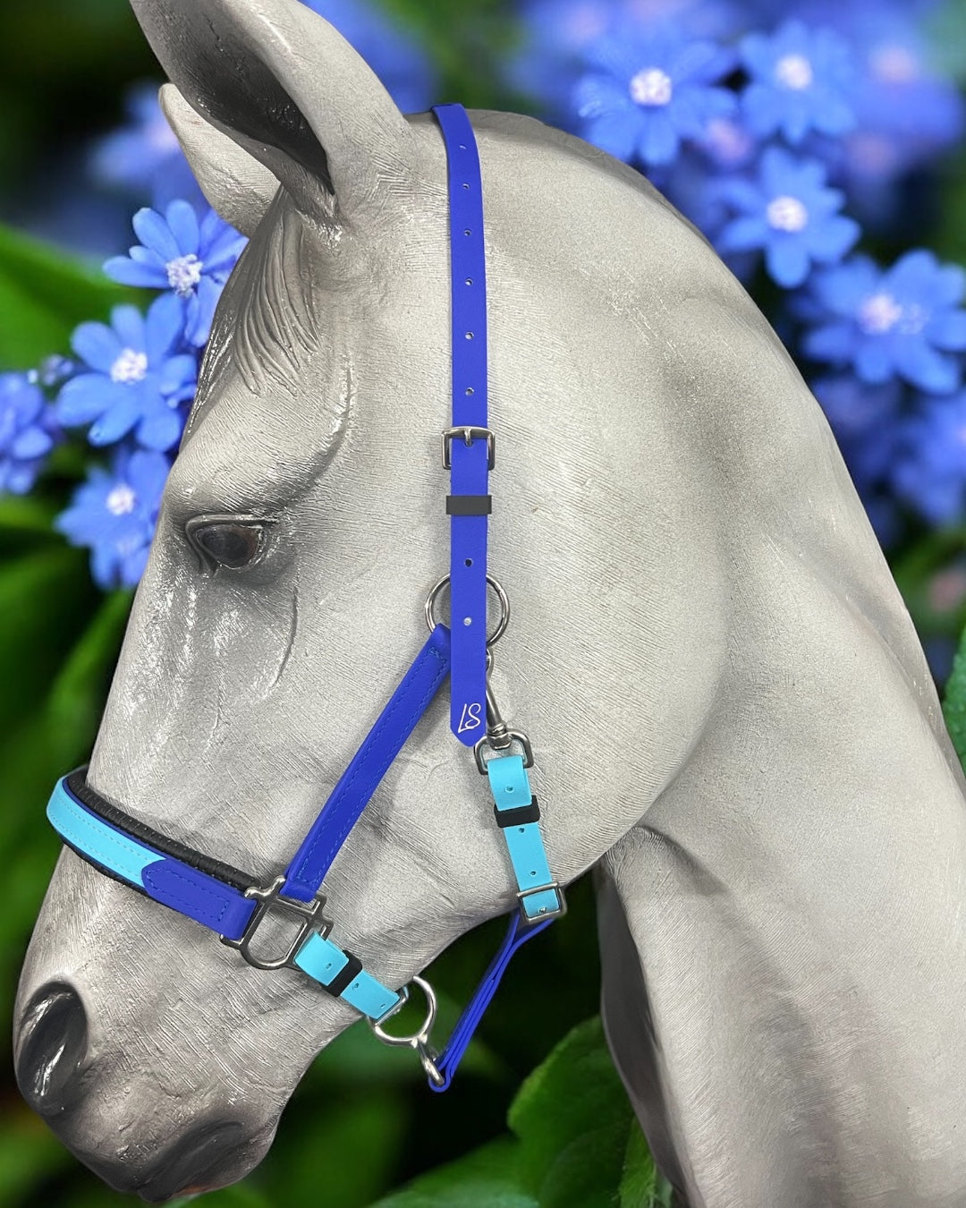 A close-up of a gray horse wearing an LS Equestrian LS Deux 2 Part Bridle Padded, designed from Beta BioThane in rich blue and cyan. The background showcases vibrant blue flowers and lush green leaves, creating a colorful and natural setting.