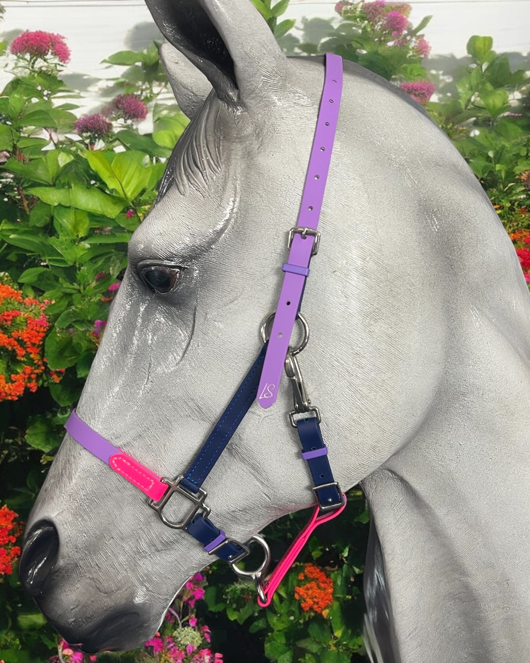 A gray horse adorned with a vibrant LS BioThane®️ Deux 2 Part Bridle from LS Equestrian—customized with pink, purple, and blue straps—stands gracefully. The background is filled with lush green foliage and vivid orange and pink flowers.