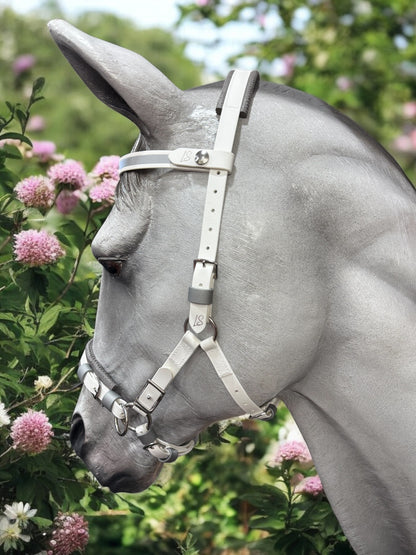 A gray horse with an LS Equestrian Premium Comfort and Control Hackamore Bridle and Jowl Strap stands among pink and white flowers in a lush garden. The setting is serene, filled with green foliage.