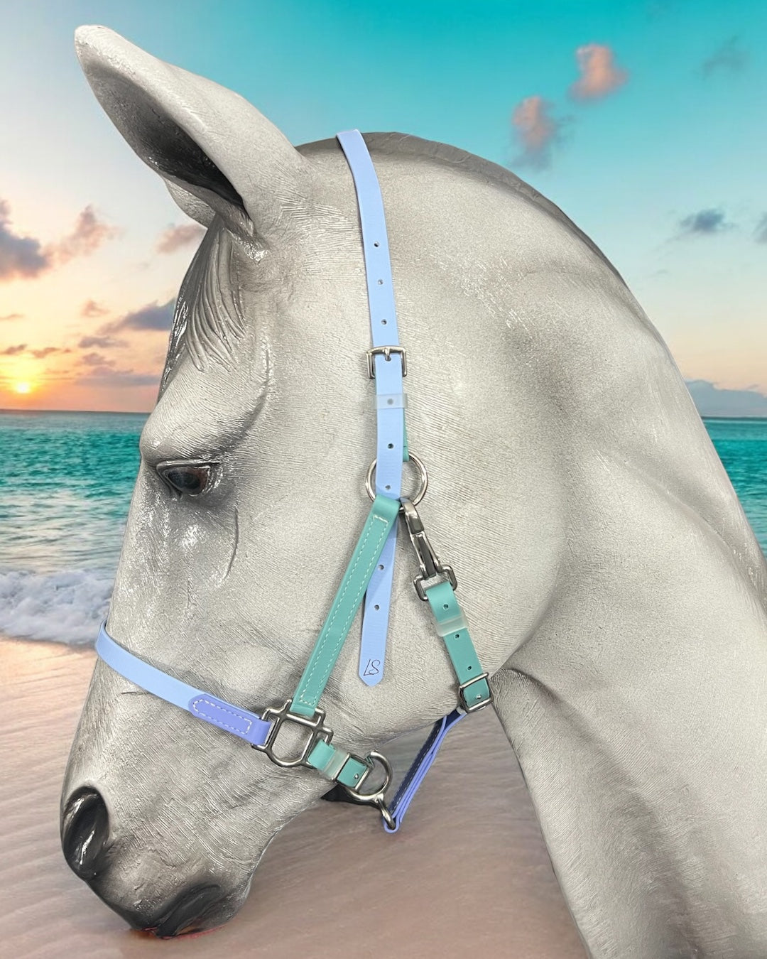 Close-up of a gray horse adorned with a pastel blue and green LS BioThane®️ Deux 2 Part Bridle from LS Equestrian, standing against a beach backdrop. The ocean is calm with a colorful sunset sky featuring pink, orange, and purple hues, perfectly complementing the custom-designed bridle that's handmade in New Zealand.