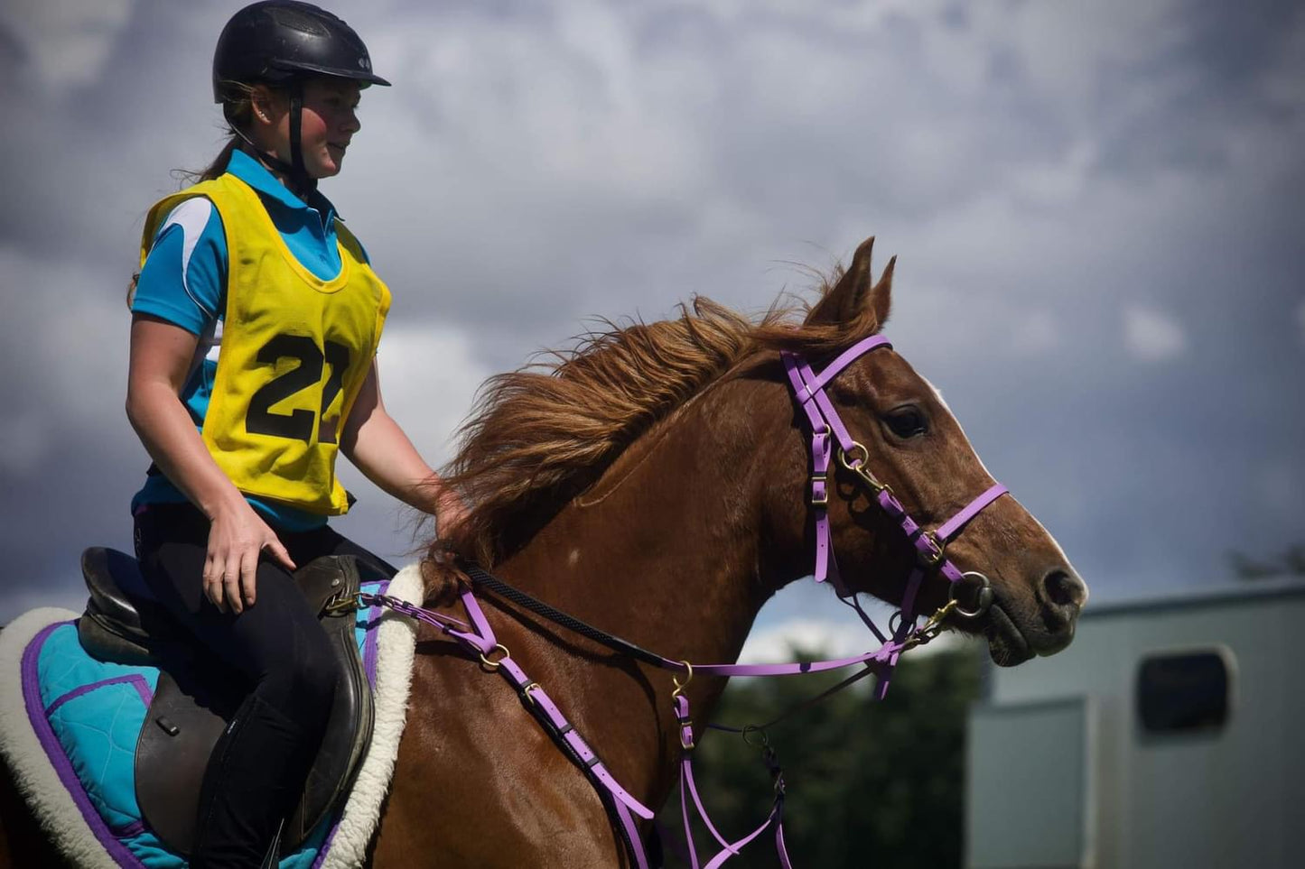 A person in a helmet and a yellow vest rides a brown horse outfitted with the LS Enduro bridle from LS Equestrian, featuring custom design stainless steel fittings. The horse's mane flows in the wind as the rider, dressed in black pants and a blue shirt, relishes the backdrop of partly cloudy skies.