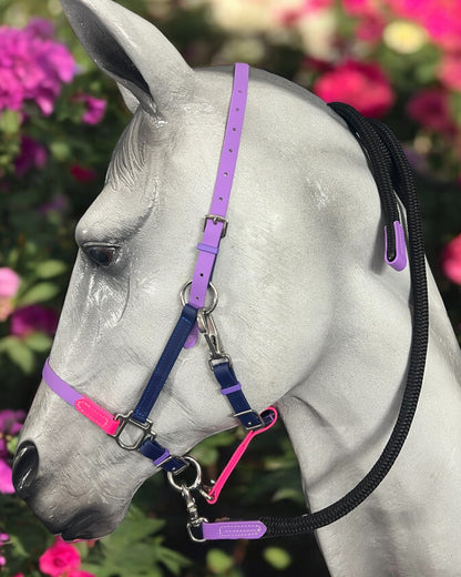 A white statue of a horse head is decorated with an LS Equestrian BioThane®️ Deux 2 Part Bridle, custom-made in New Zealand, showcasing lively hues of purple, pink, and black. The backdrop flourishes with vibrant pink flowers and rich green foliage.