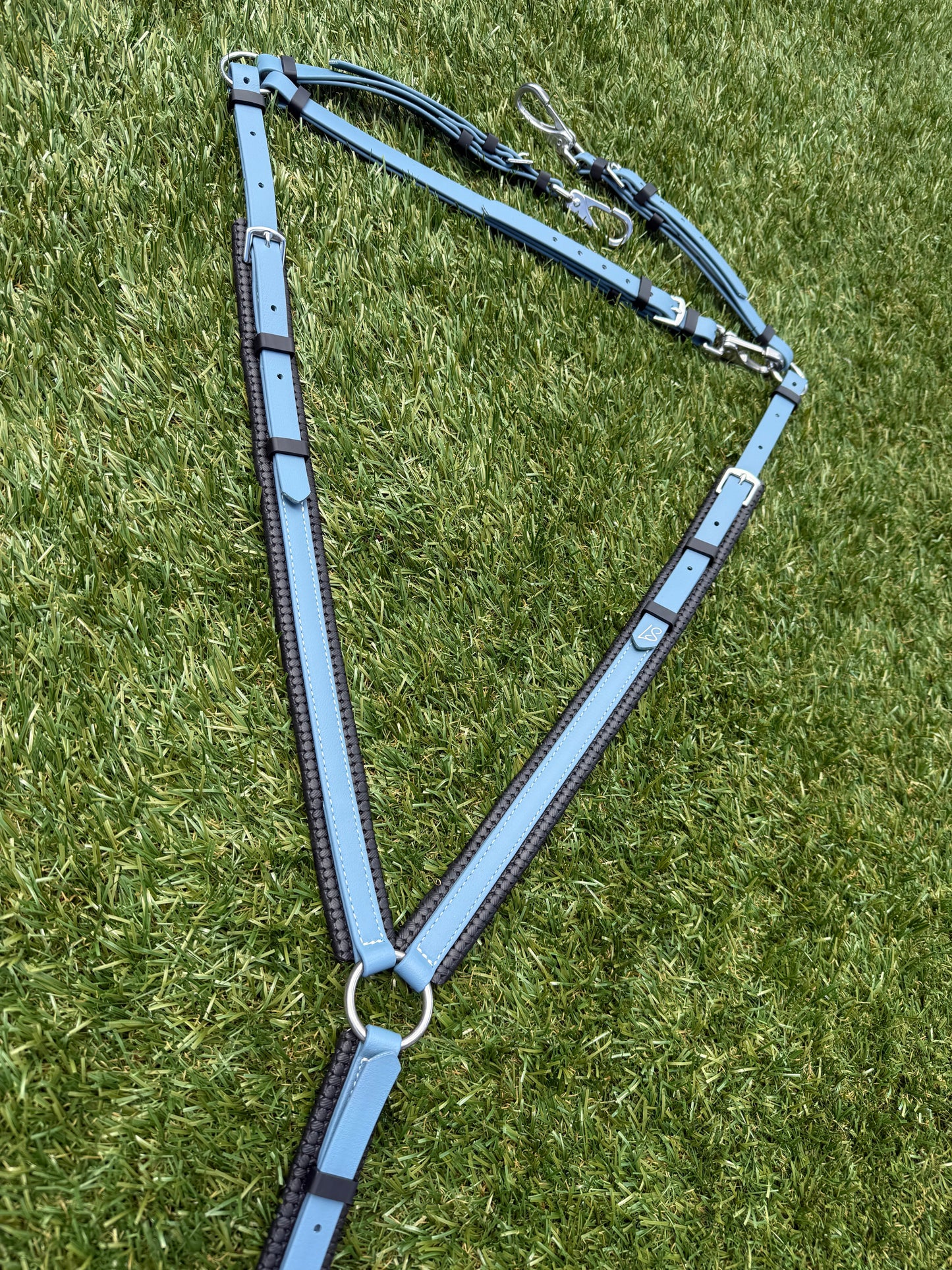 The LS Padded Breastplate - Petrol Blue by LS Equestrian NZ, featuring a Petrol Blue and black design with silver hardware, rests on green grass in a triangular shape. The Beta BioThane leather straps are neatly arranged, showcasing the metal rings and clips clearly.