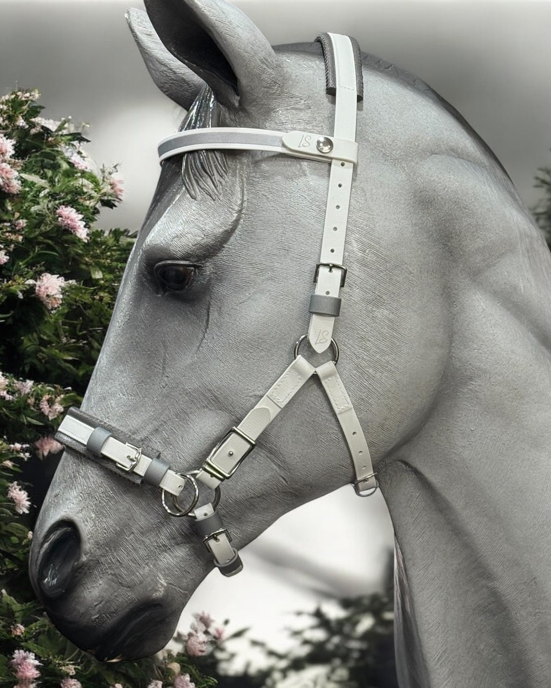 A gray horse, outfitted in an LS Hackamore Bridle with Jowl Strap by LS Equestrian, stands amidst green foliage and pink flowers. The horse faces left, highlighting the bridle's sleek design.