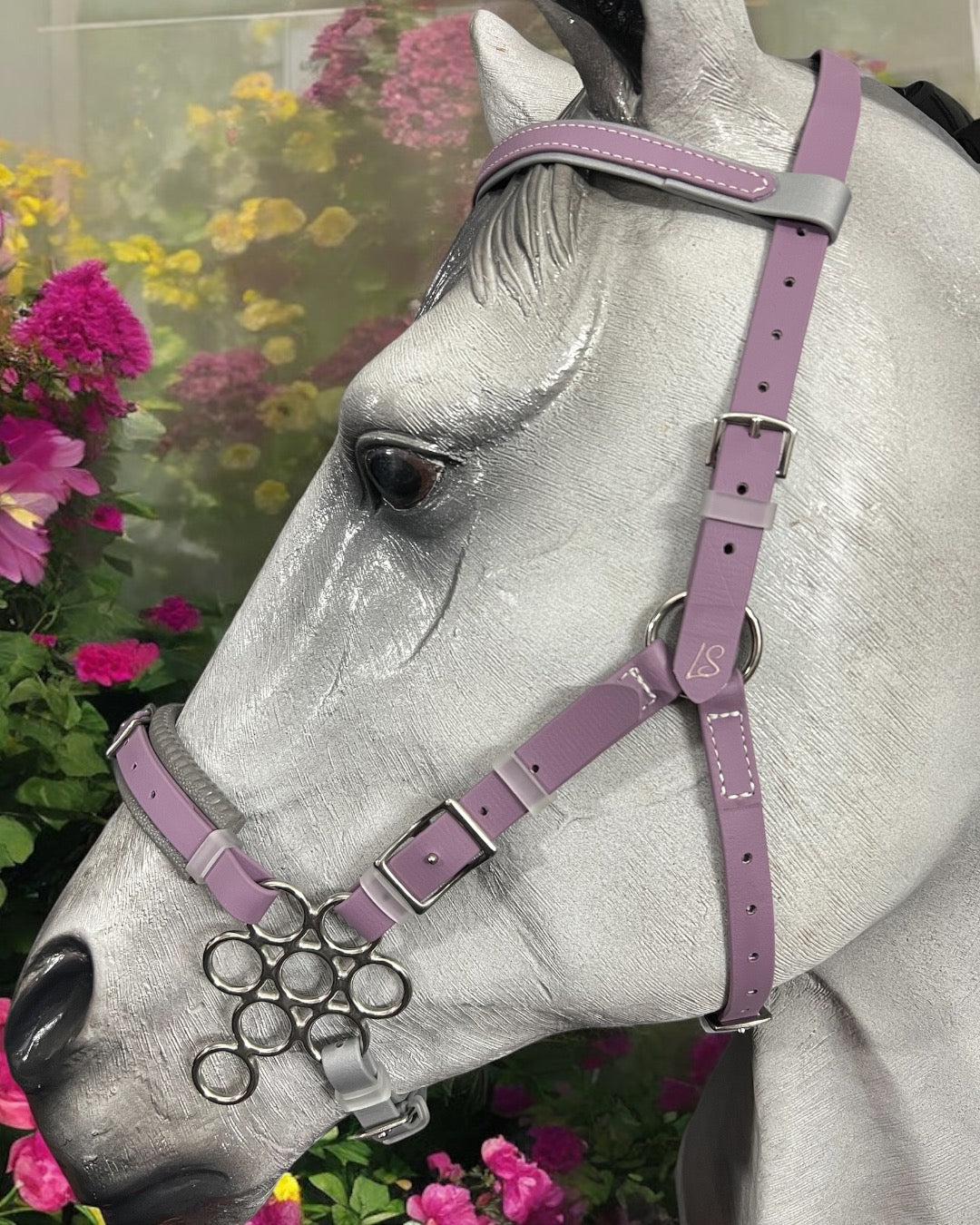 A gray horse mannequin elegantly showcases an LS Hackamore Bridle with Jowl Strap for premium comfort and control by LS Equestrian, crafted in lavender BioThane® with decorative rings against vibrant pink and purple flowers.