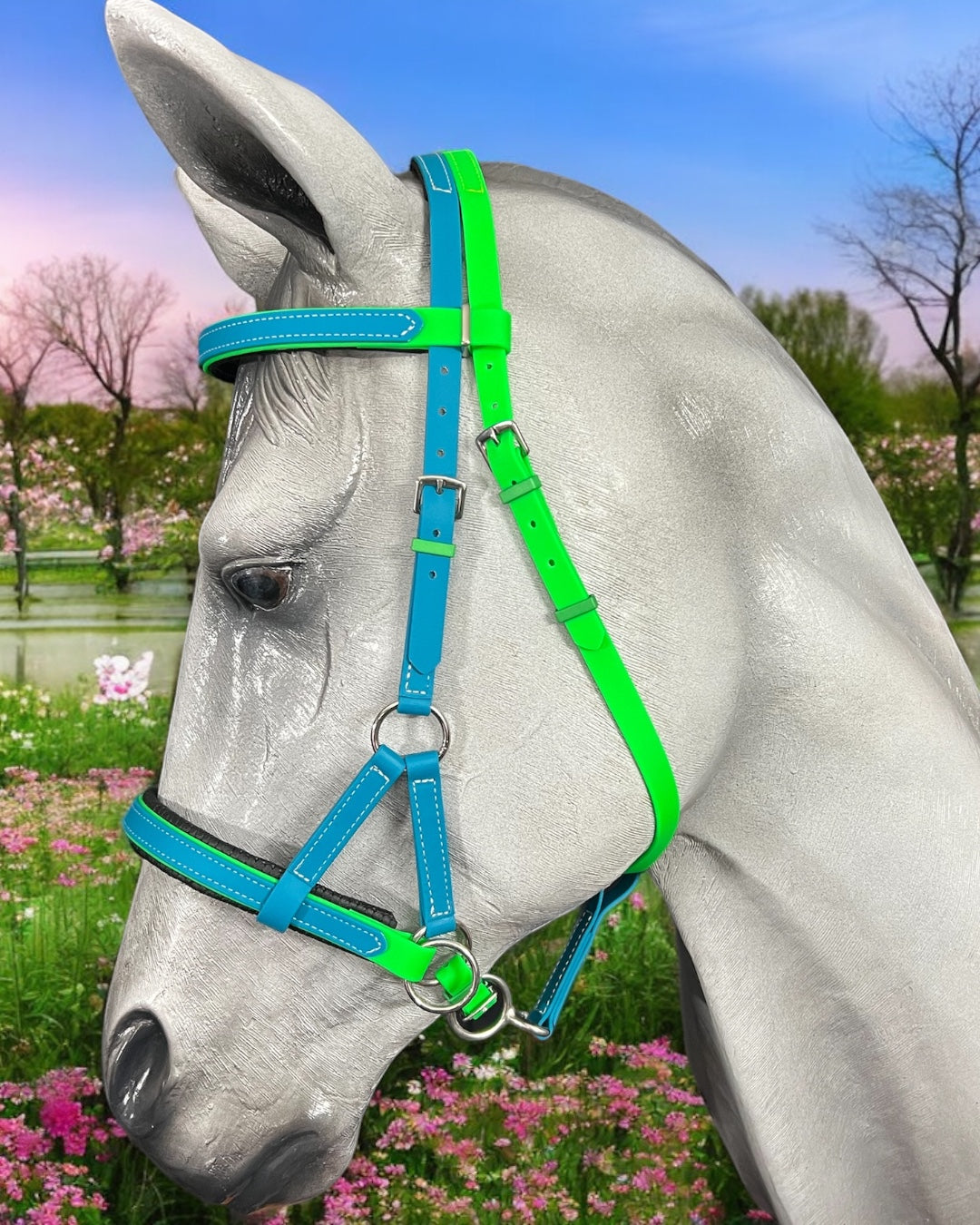 A gray horse sports the LS Equestrian SidePull Bitless Bridle Option 2 in vibrant green and blue amid blooming pink and white flowers, with a serene spring landscape of trees and a pond under a blue sky.