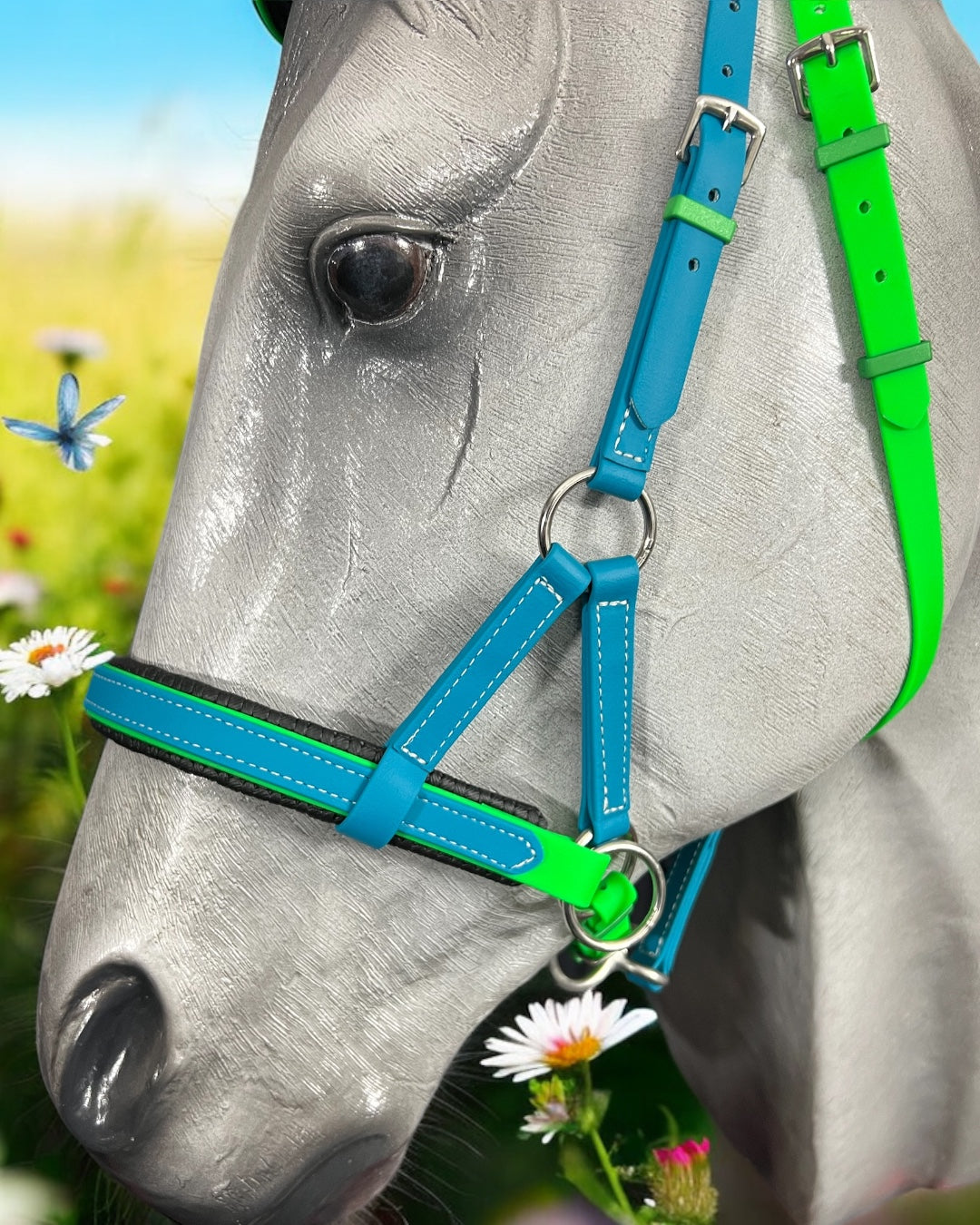 A gray horse statue wears a custom blue and green SidePull Bitless Bridle Option 2 from LS Equestrian, surrounded by white daisies and a small blue butterfly in the lush grass background.