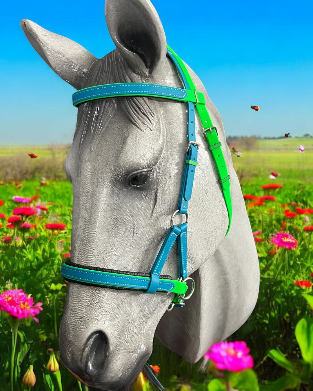 A statue of a horse head stands in vibrant pink flowers, adorned with a bright blue and green SidePull Bitless Bridle Option 2 by LS Equestrian. Butterflies flutter gracefully against the clear blue sky, completing the enchanting scene.