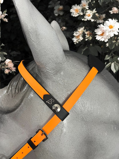 A close-up of a horse sculpture showcases the LS Equestrian Multi Bridle Option 3 - Design your Own, in a vibrant bright orange with a sturdy metal clasp. The scene is beautifully set against a backdrop of white flowers and lush green foliage, creating an eye-catching contrast.