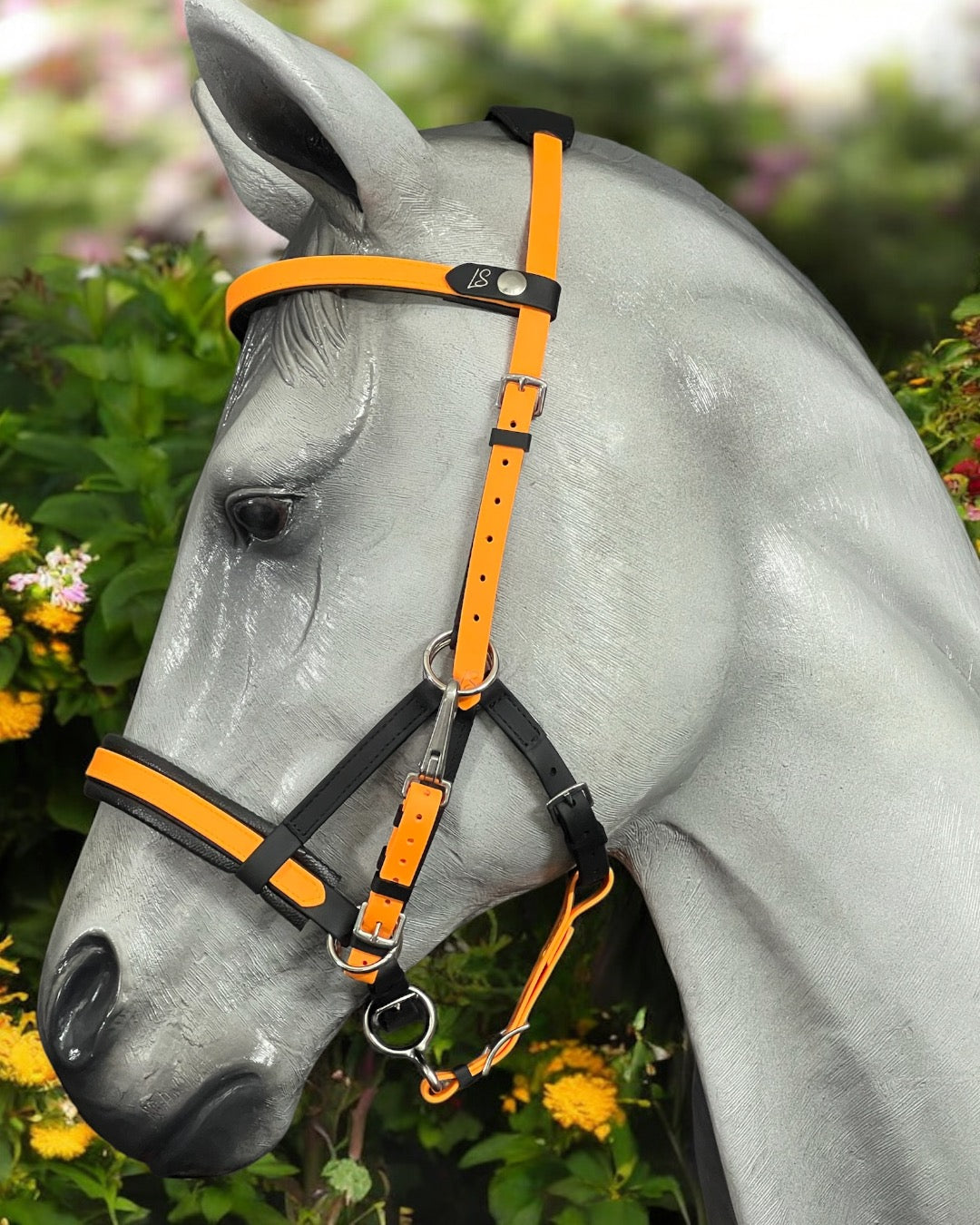 A gray horse, wearing a striking custom orange and black halter from LS Equestrian's Multi Bridle Option 3 - Design your Own line, stands amidst lush greenery and colorful flowers. Its sleek appearance is accentuated as it gazes to the side, framed by a softly blurred background.