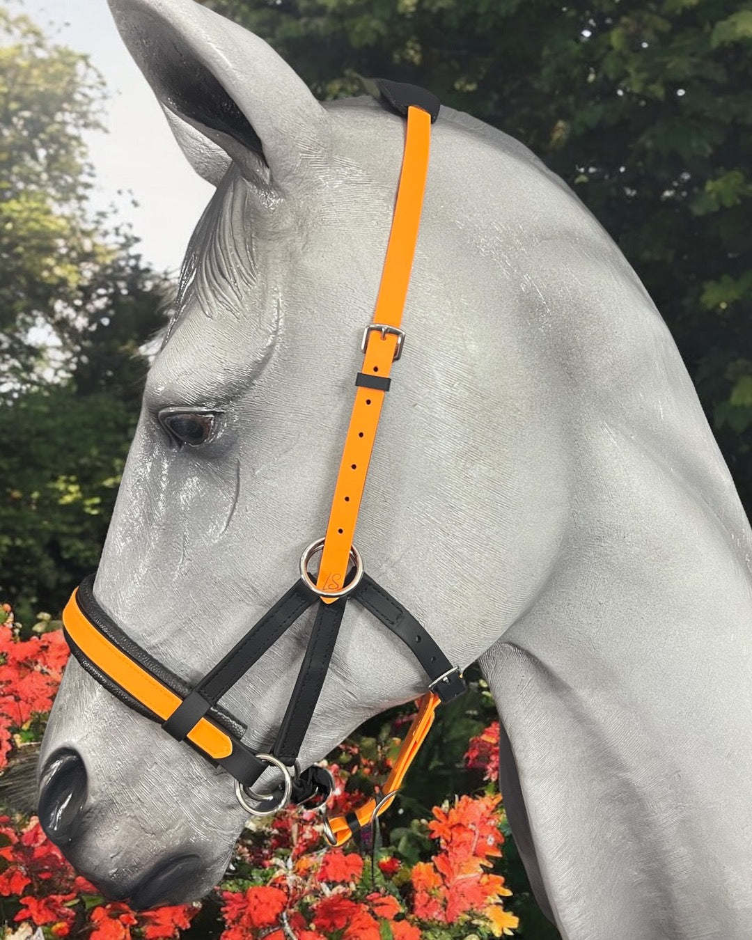 A gray horse, adorned in an LS Equestrian Multi Bridle Option 3 - Design your Own halter in black and orange BioThane, stands near vibrant red flowers. Lush green foliage provides a serene backdrop.