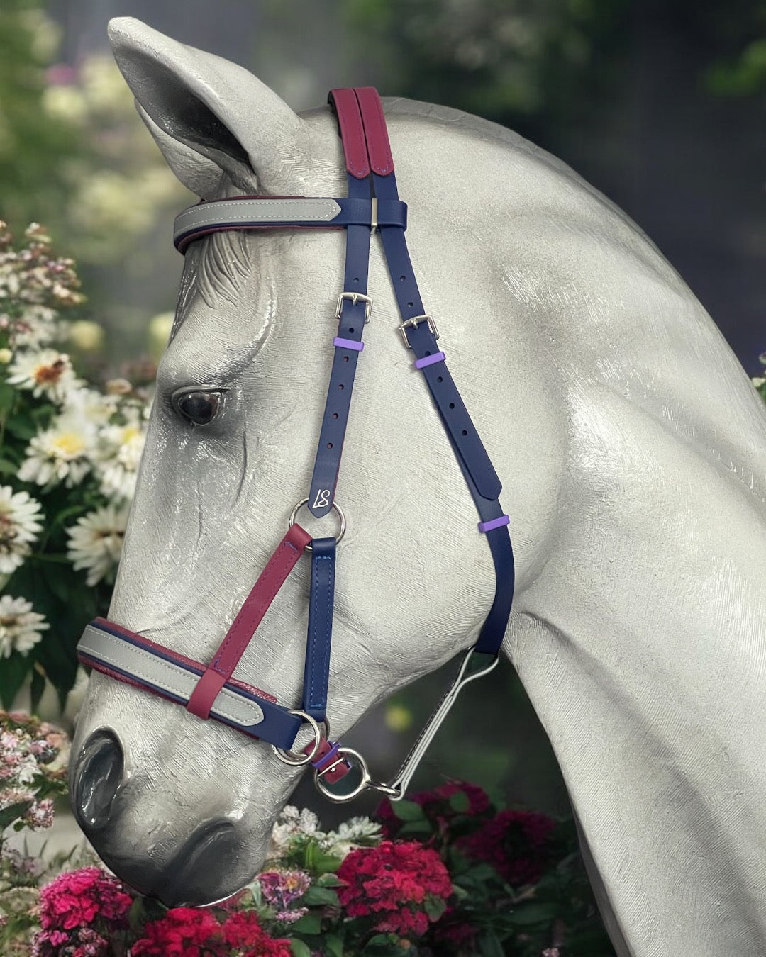 A horse wearing an LS Equestrian SidePull Bitless Bridle Option 2 with customizable red, blue, and purple straps stands against a backdrop of greenery and white flowers.