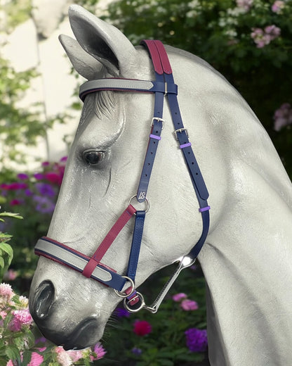 A gray horse stands gracefully in front of lush green and pink foliage, wearing an LS Equestrian SidePull Bitless Bridle Option 2 featuring vibrant red, blue, and purple custom-made BioThane®️ straps.