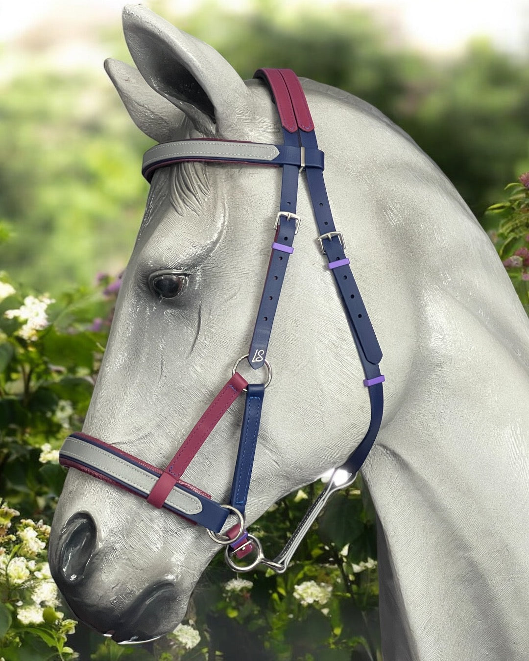 A gray horse stands elegantly before green foliage and white flowers, wearing an LS Equestrian SidePull Bitless Bridle with navy blue, maroon, and light gray BioThane®️ straps.