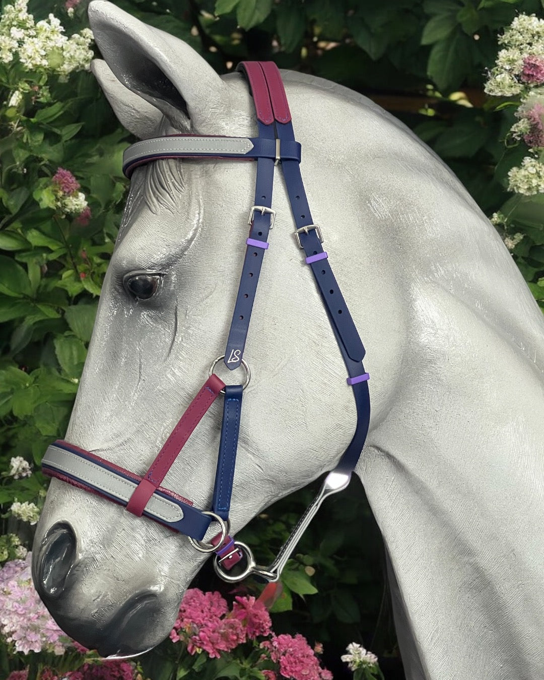 A silver horse mannequin is adorned with an LS Equestrian SidePull Bitless Bridle Option 2, designed with blue, purple, and pink BioThane®️ straps, set against a vibrant green backdrop of blooming flowers.