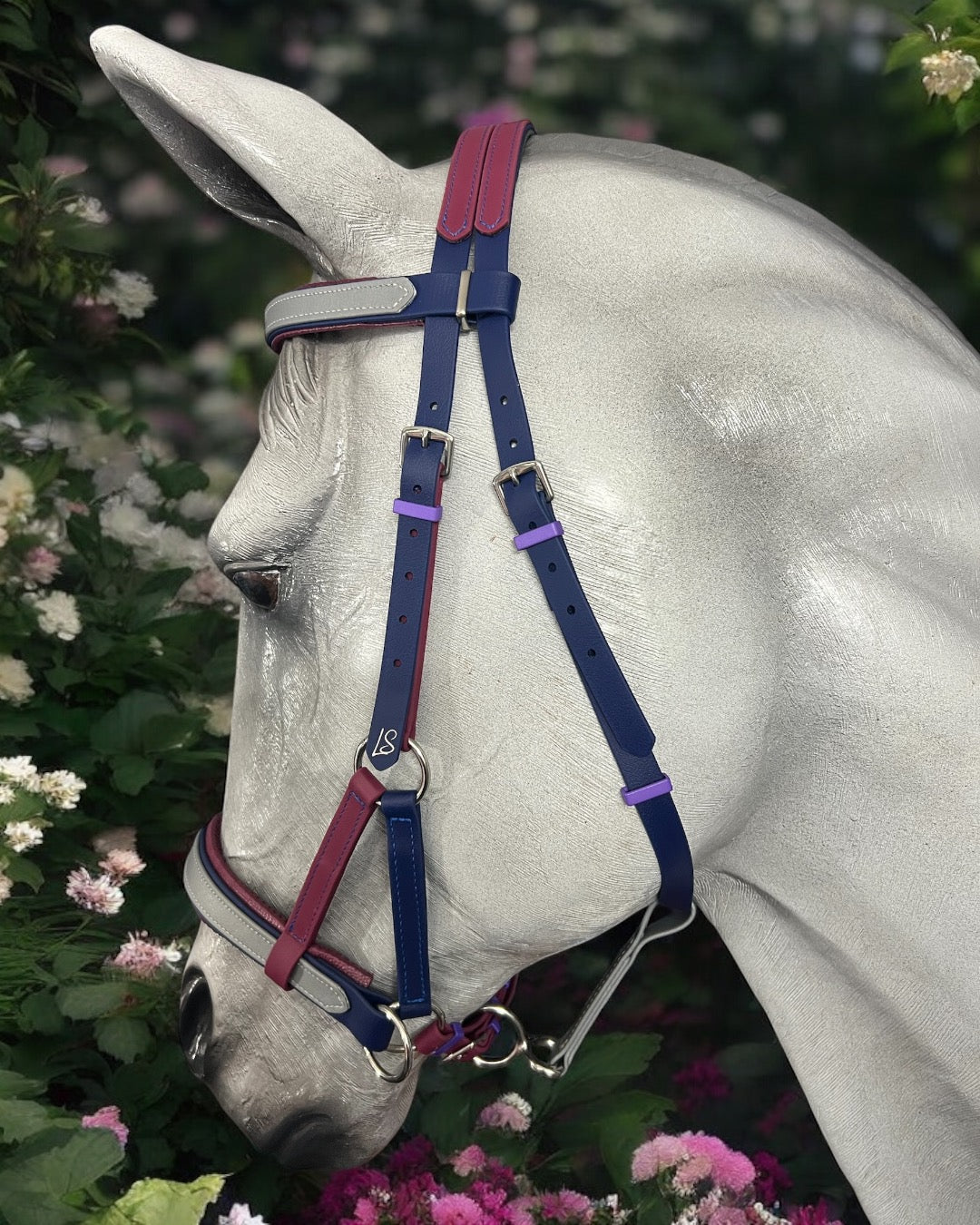 A white horse adorned with a custom-made LS Equestrian SidePull Bitless Bridle Option 2, featuring blue, red, and contrasting purple accents, stands amidst lush greenery and pink flowers, adding a colorful touch to the elegant scene.