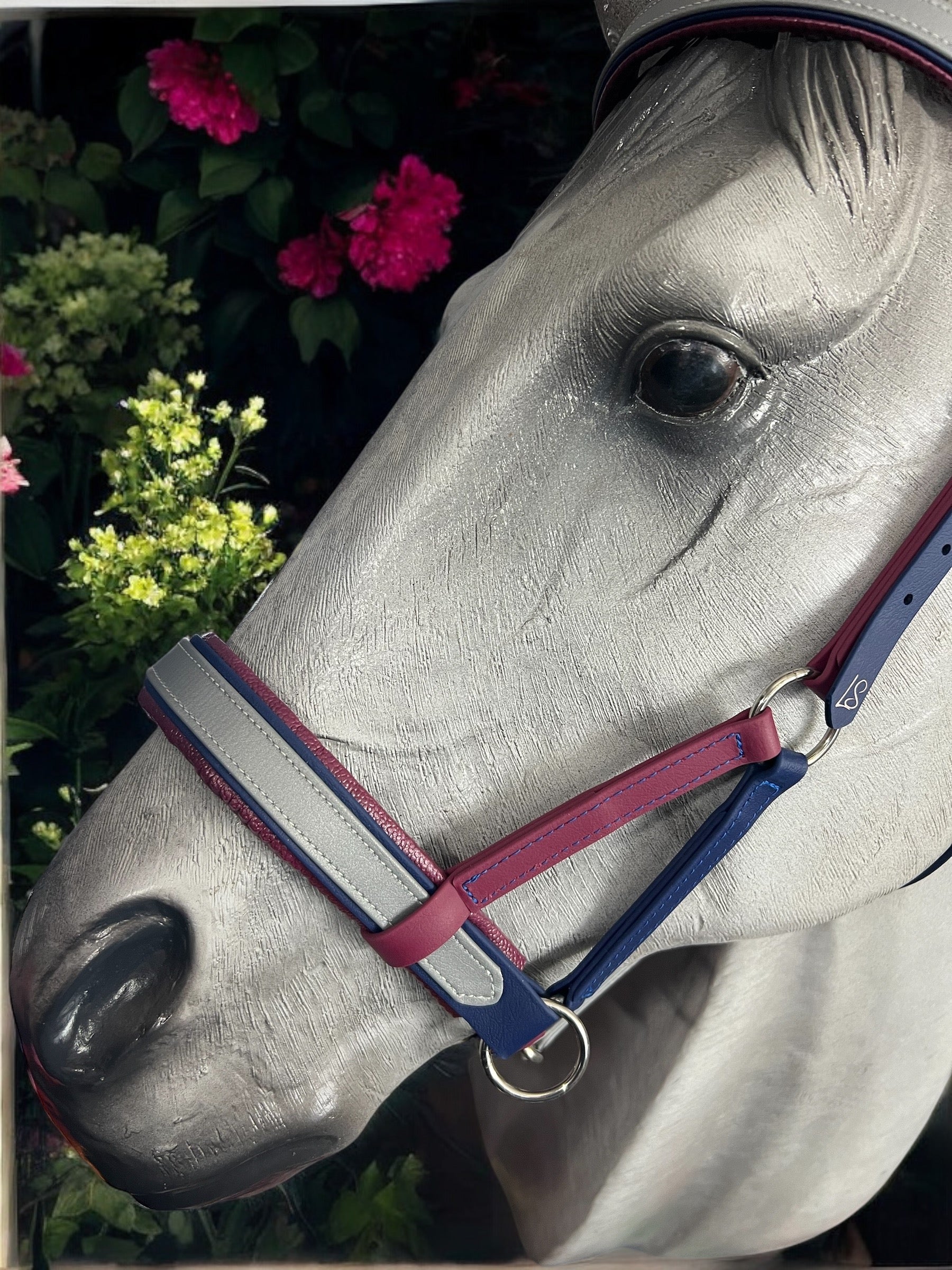 A gray horse sculpture features a custom LS Equestrian SidePull Bitless Bridle Option 2 with red and blue straps, set against a backdrop of lush greenery and pink flowers.