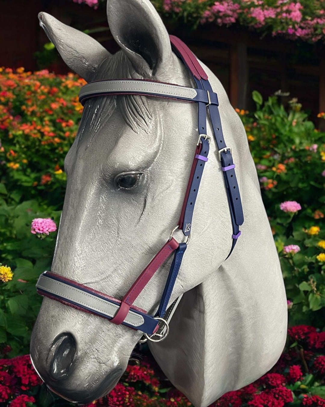 A gray horse statue with a vibrant LS Equestrian SidePull Bitless Bridle stands before a lush garden, showcasing pink and yellow flowers amidst abundant green foliage.